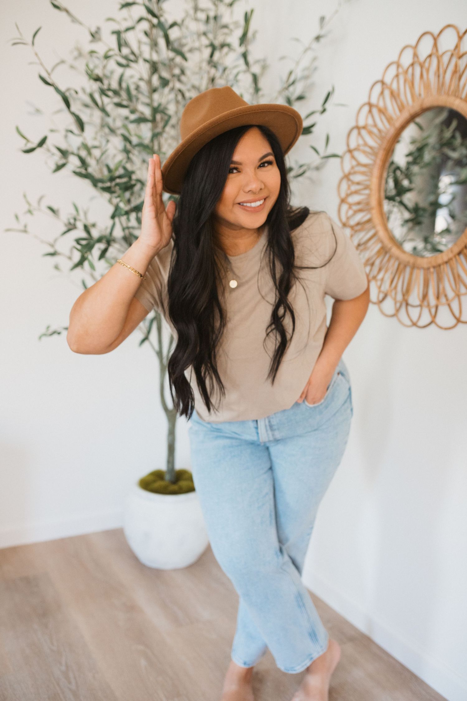 Lifestyle fashion photoshoot featuring light beige sweater, blue jeans and brown hat against white wall with greenery.