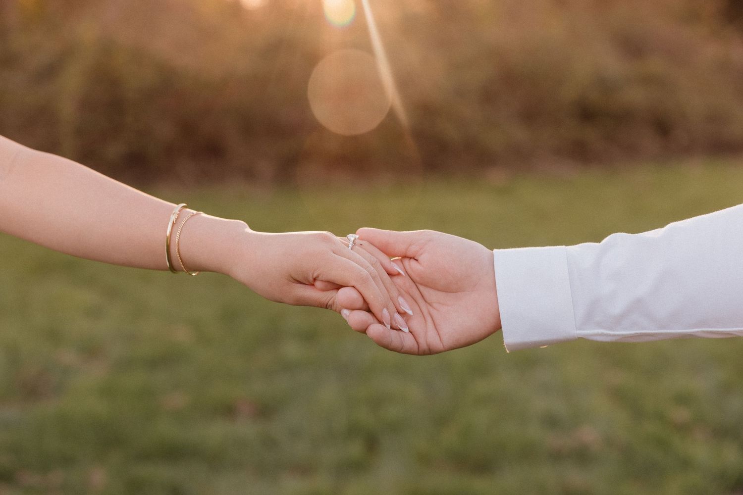 Two people gently holding hands outdoors at golden hour, one wearing a gold bracelet and engagement ring.