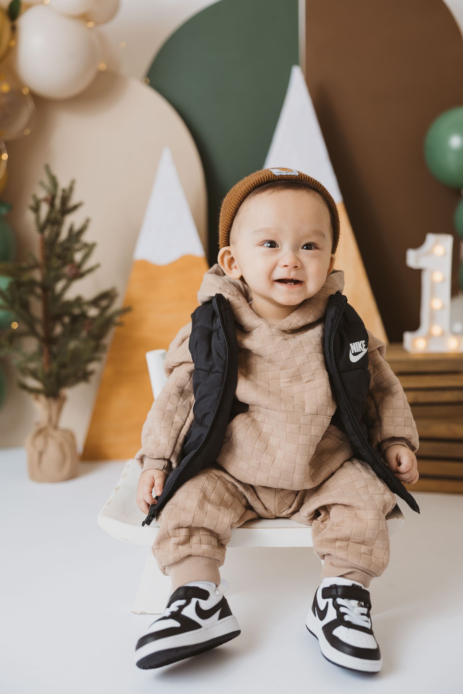 Baby wearing beige outfit and sneakers sitting against festive balloon backdrop.