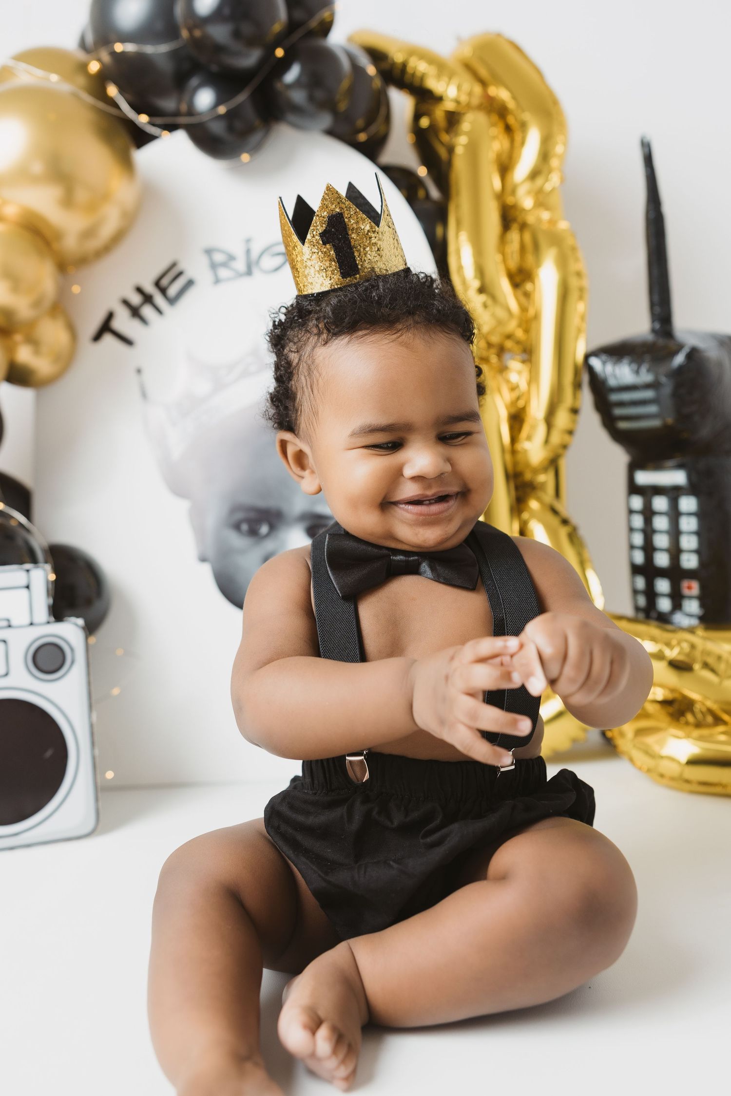Smiling baby in black outfit and gold crown celebrates first birthday with balloons and retro decorations.