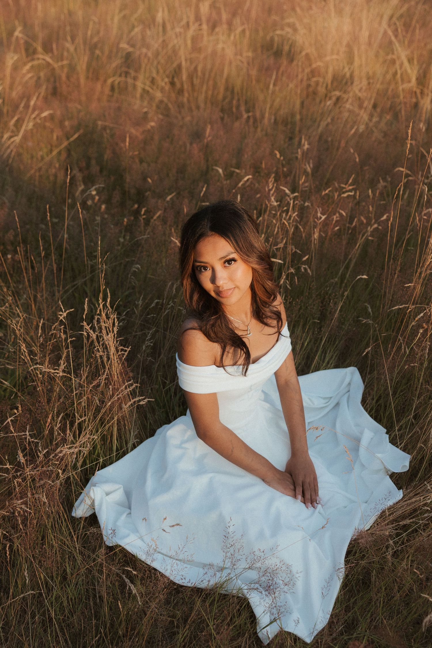Young woman in white off-shoulder gown sitting gracefully in golden tall grass field during sunset.
