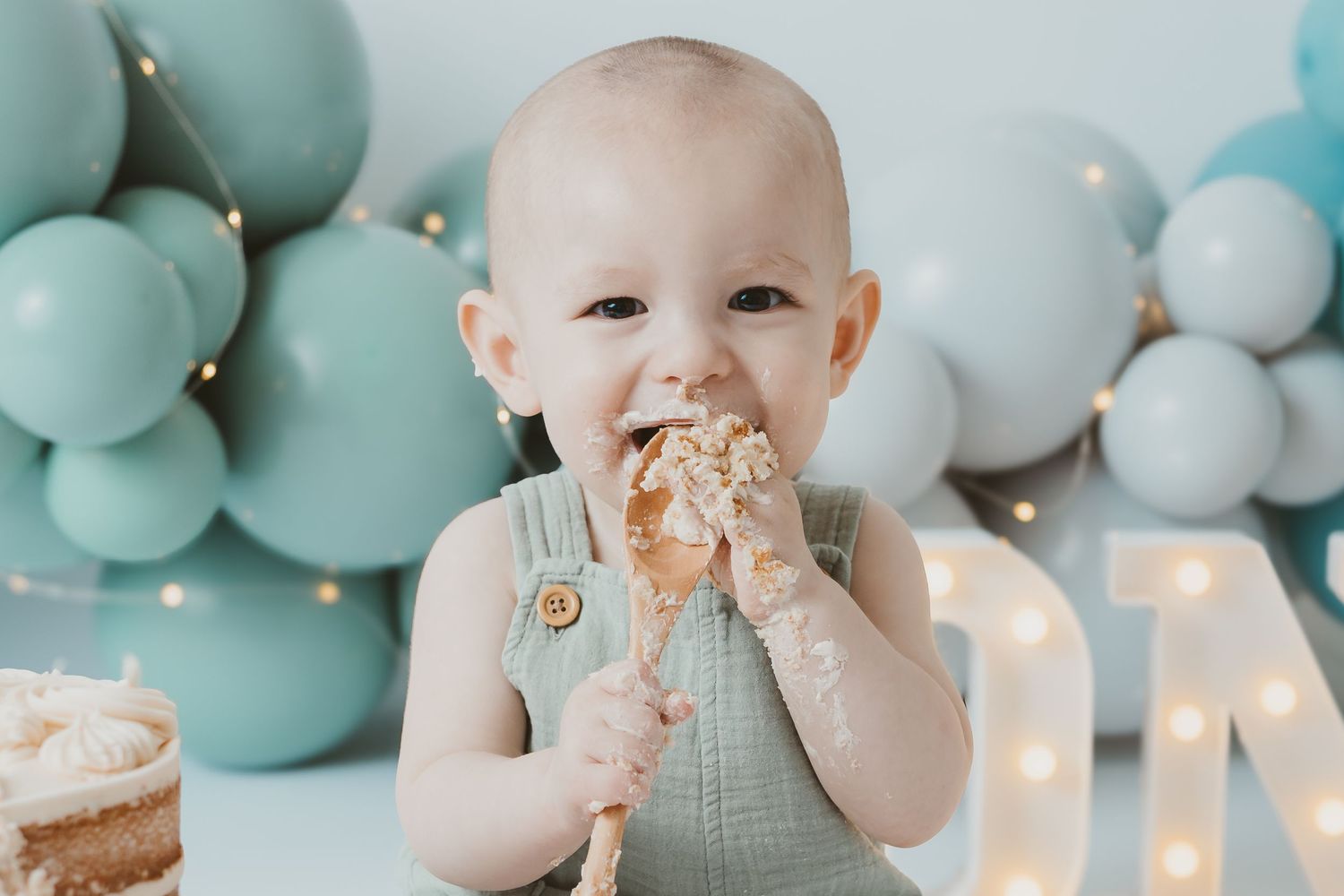 A baby in mint overalls enjoys cake during first birthday photoshoot with blue balloons.