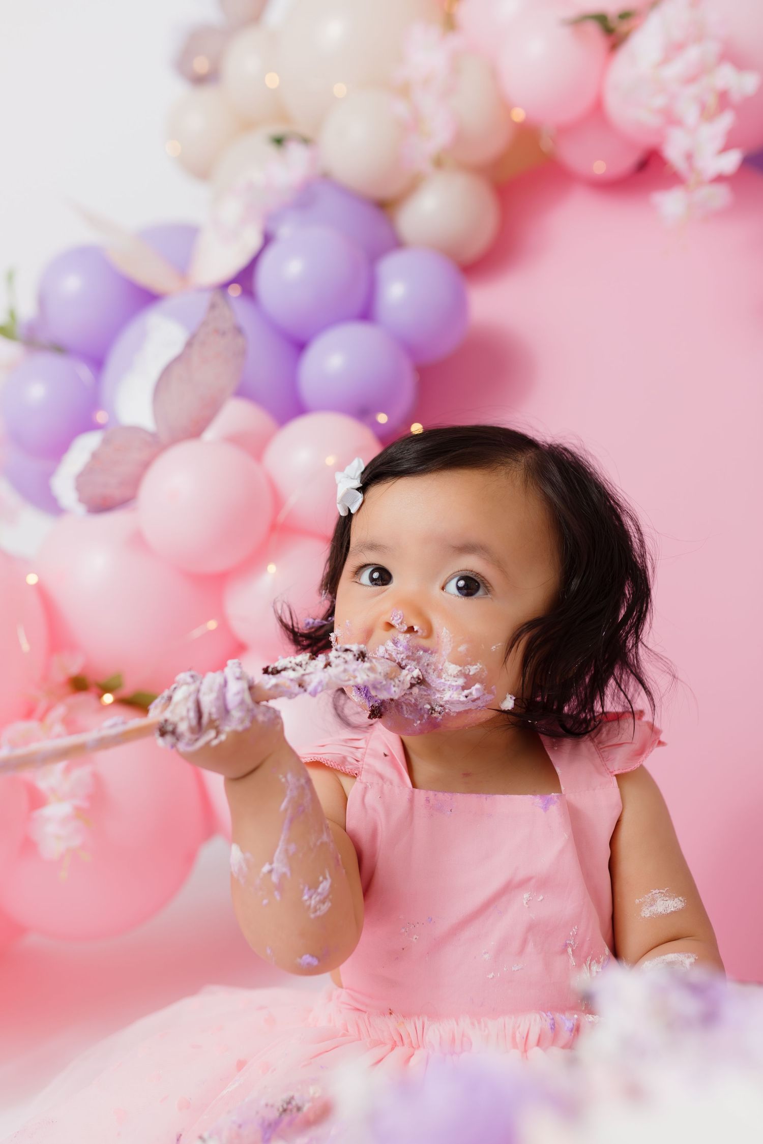 A baby in pink tulle dress plays with confetti against pastel balloon decorations.