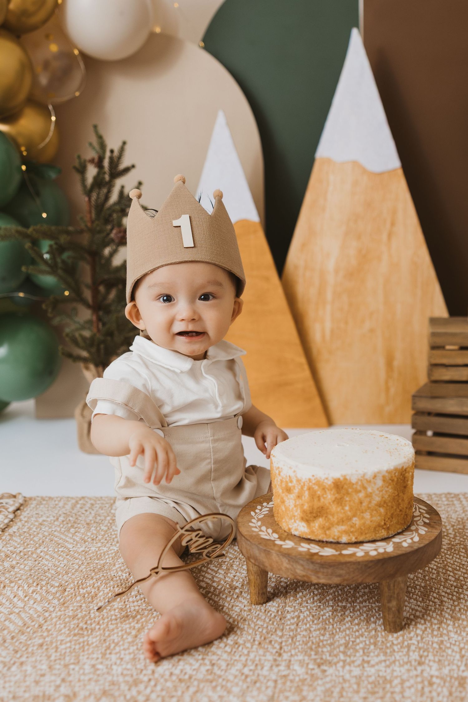 First birthday celebration setup with white and gold balloons, wooden decorations, and a white cake on a textured rug.