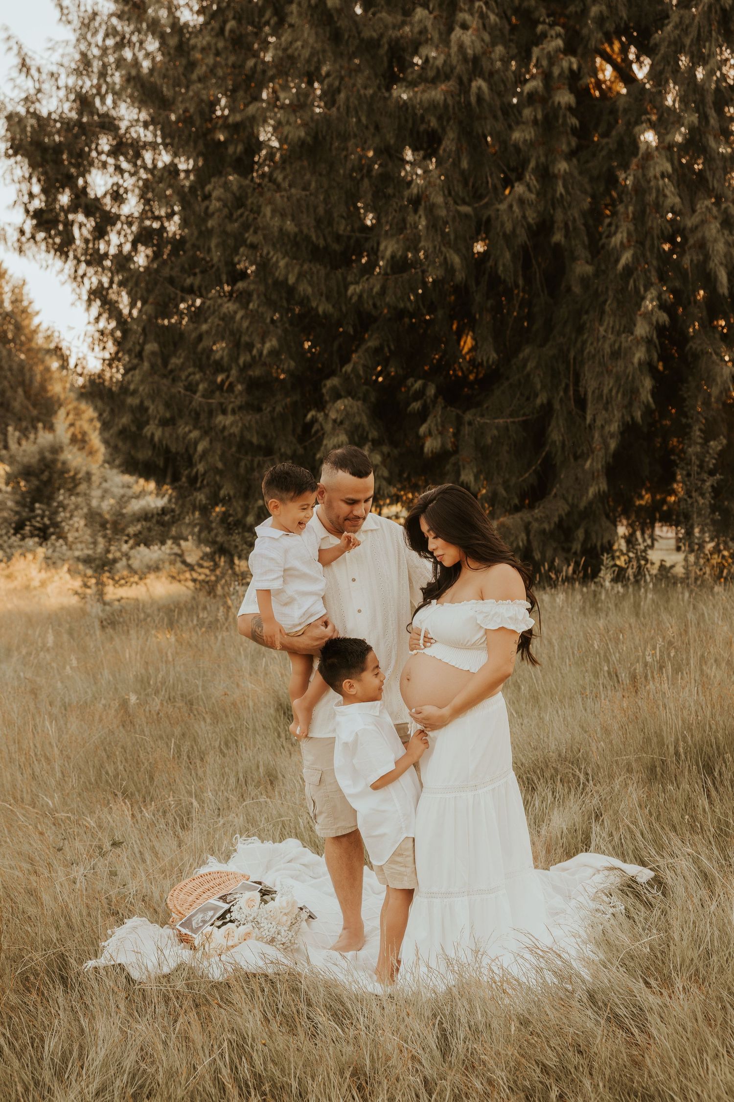 Pregnant woman in white dress poses with husband and two young sons in golden field for maternity photo.
