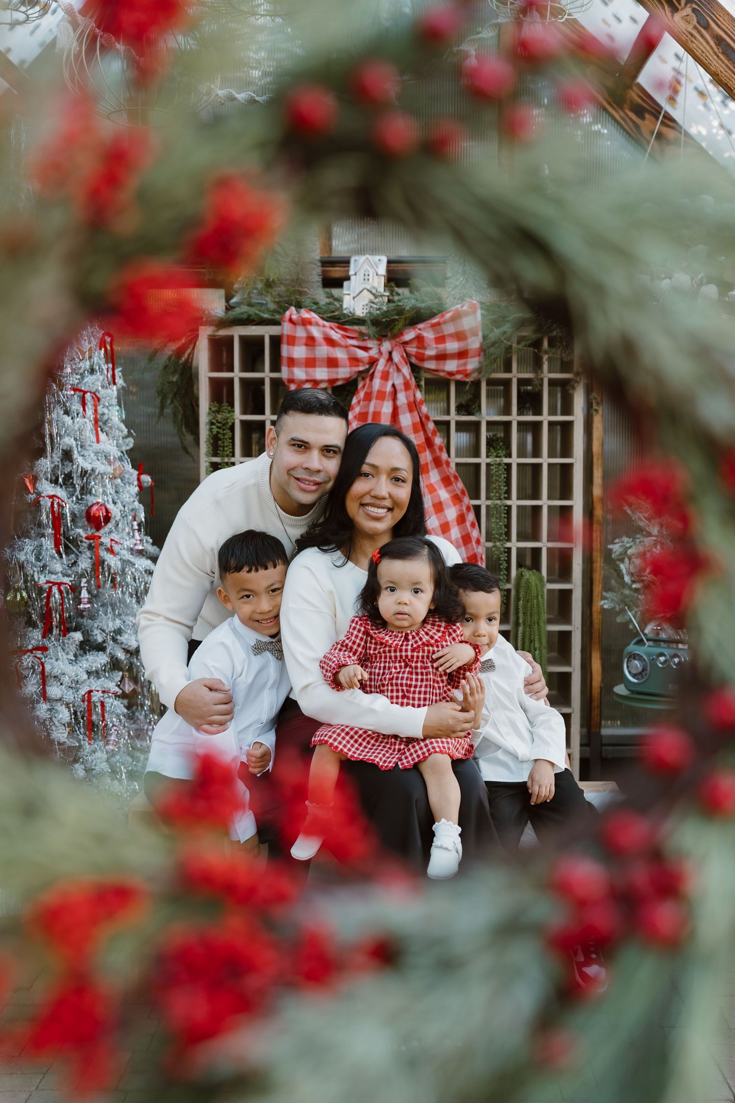 Family of four poses for a festive Christmas photo surrounded by red berries, holiday greenery, and a decorated white tree.