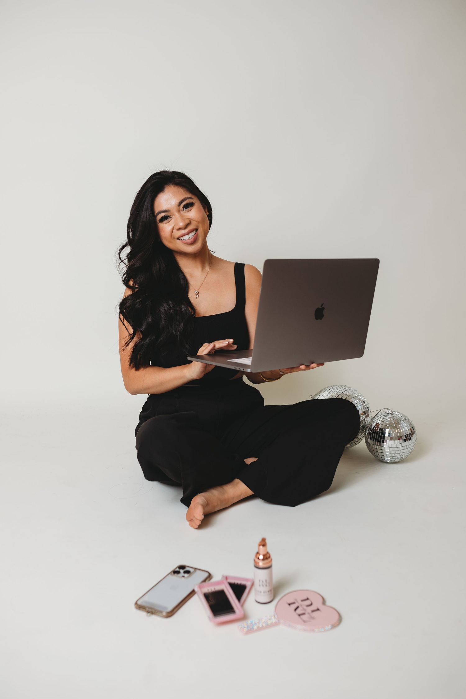 Person in black outfit working on laptop with phone and accessories on white floor.