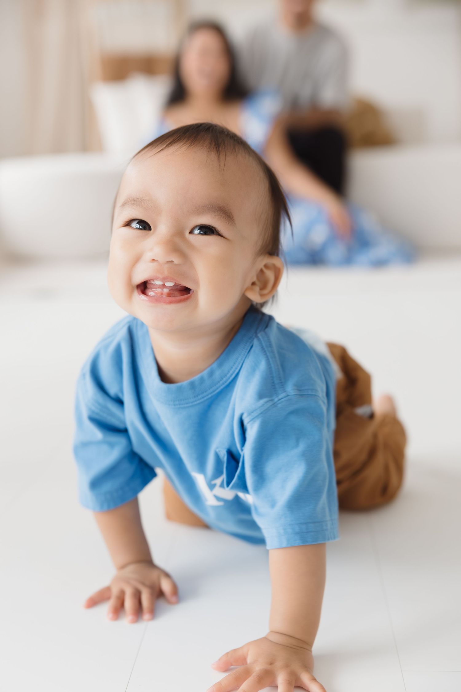 Baby in light blue shirt crawls and smiles on white surface.