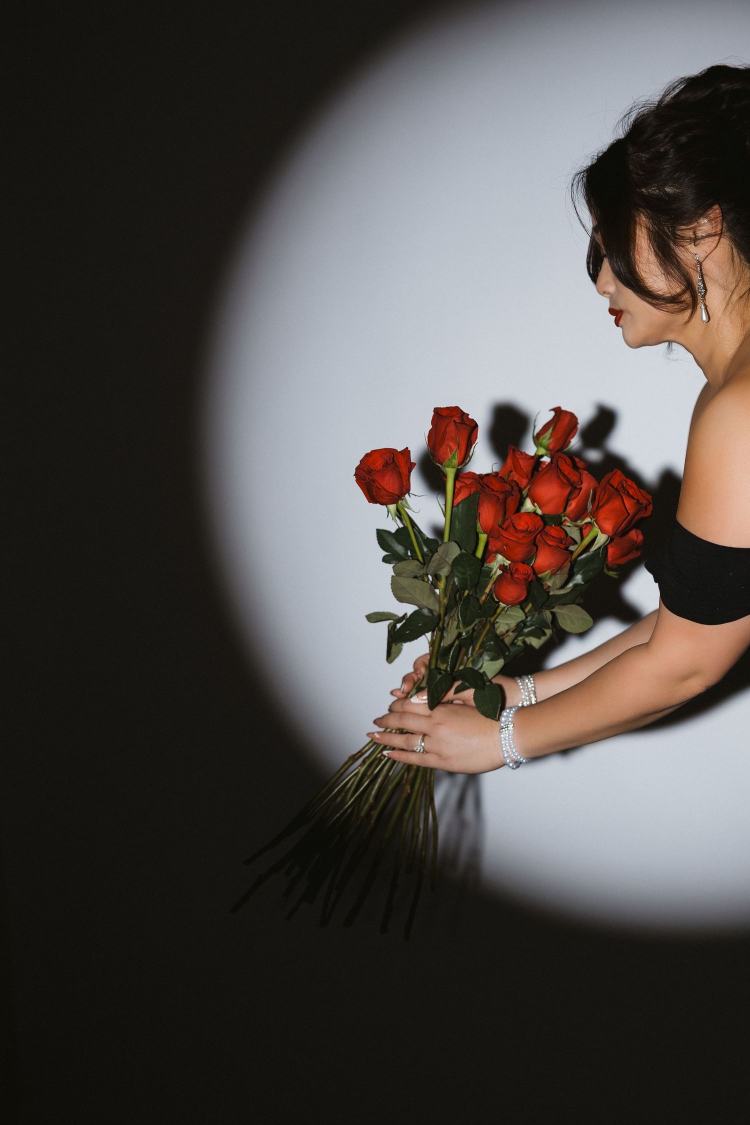 Woman in black dress holding bouquet of red roses against dramatic spotlight backdrop.