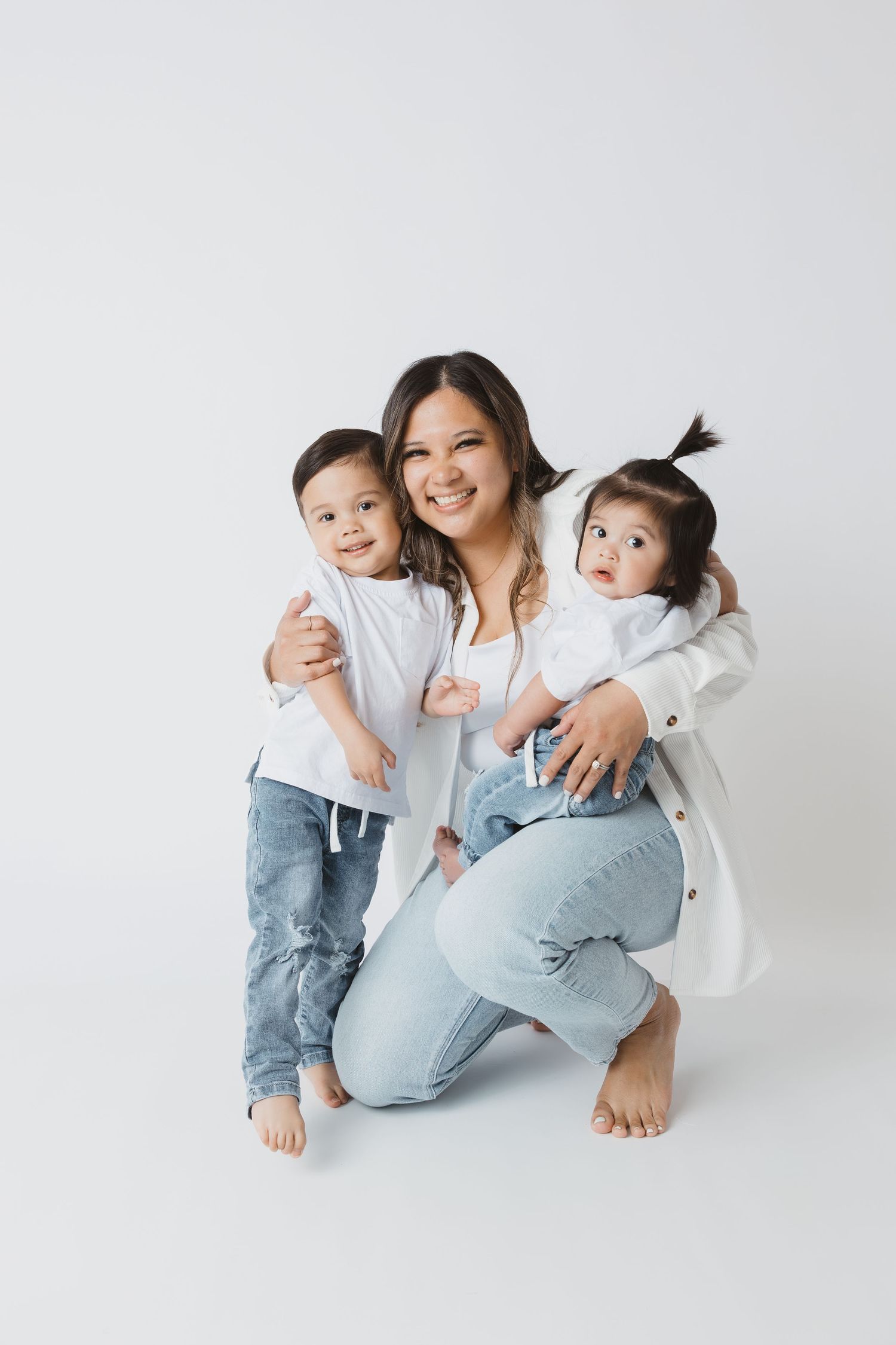 A smiling woman crouches with two young children, all wearing white tops and jeans, posing together on a white background.