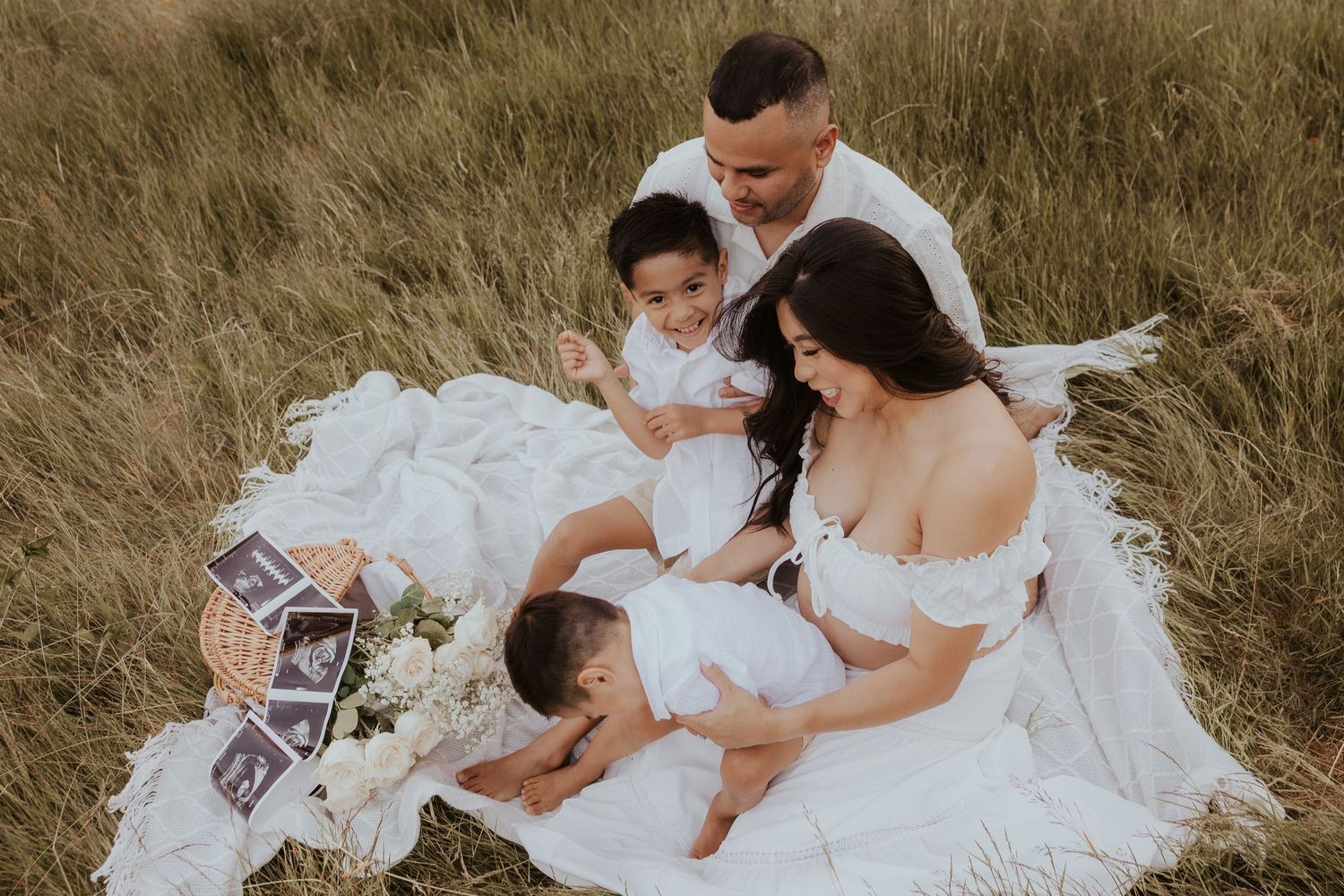Family maternity photo shoot outdoors in a field, parents and two children in white outfits on a blanket with ultrasound photos.