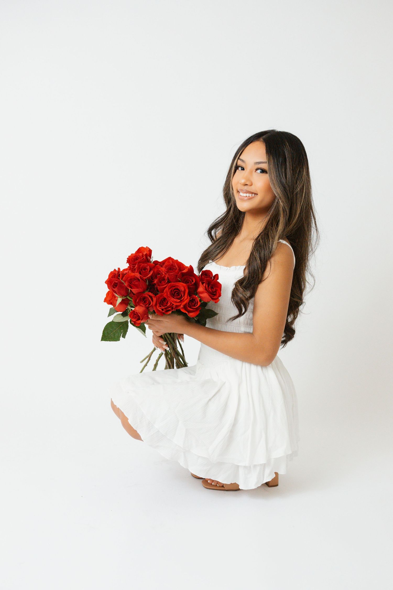 Woman in white dress poses with red roses against white backdrop in studio setting.