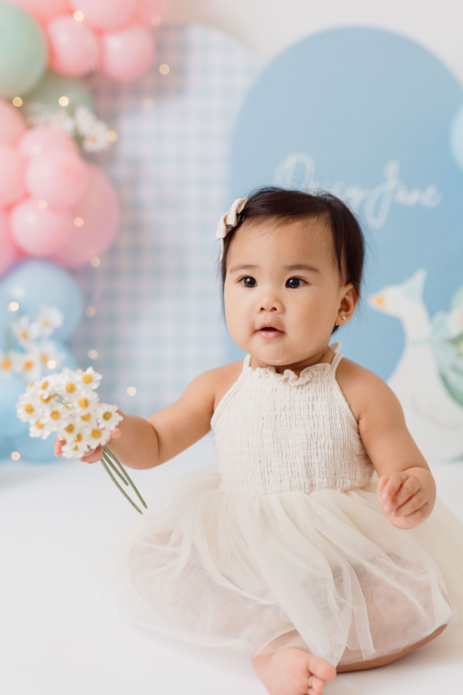 A baby in a white tulle dress poses with sparkly decorations against a pastel balloon backdrop.