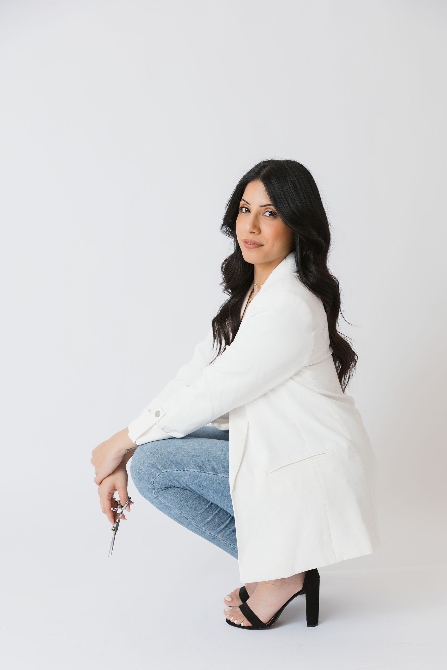 Woman in white blazer and jeans sits in minimalist studio setting.