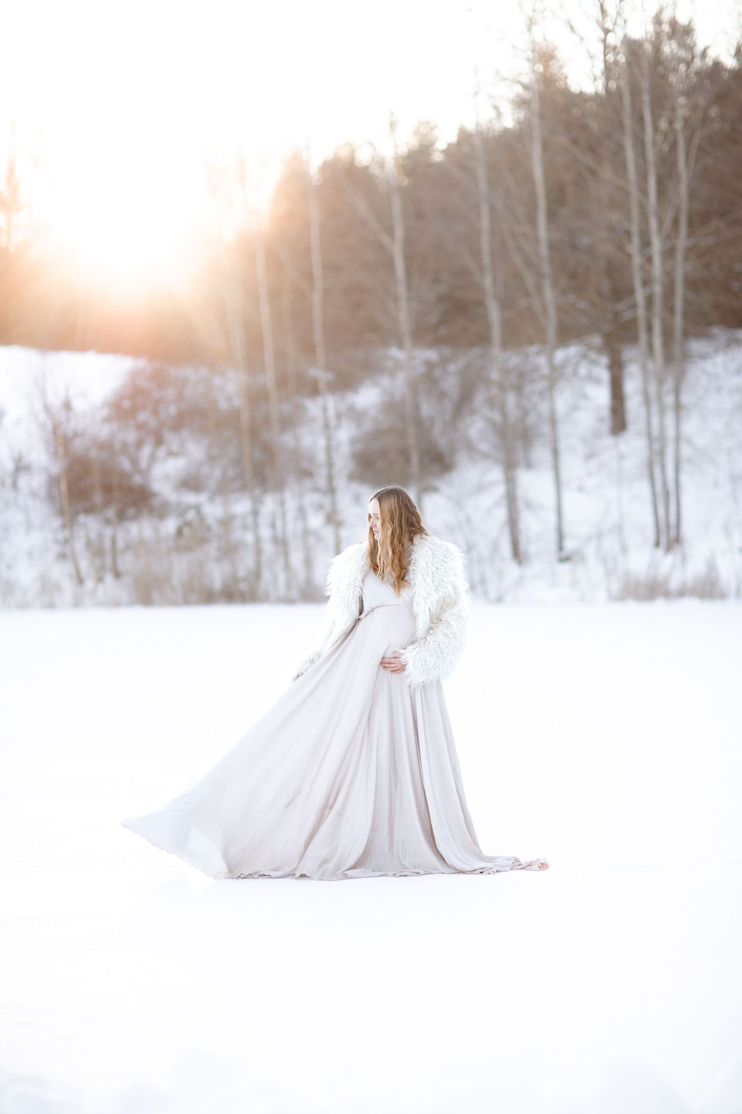 Gravidfotografering i vacker klänning i ett vinterlandskap. Mamman kollar ner mot magen och ser drömsk ut.