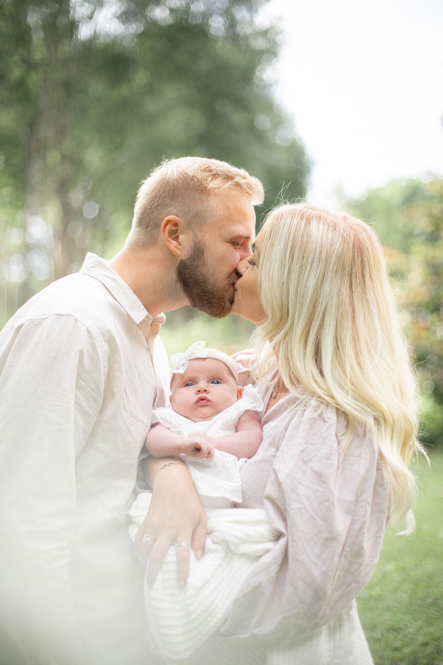 Mamma och pappa pussas med den nyfödda i famnen. Nyföddfotografering utomhus i vacker sommargrön miljö. Familjefotograf i Katrineholm.