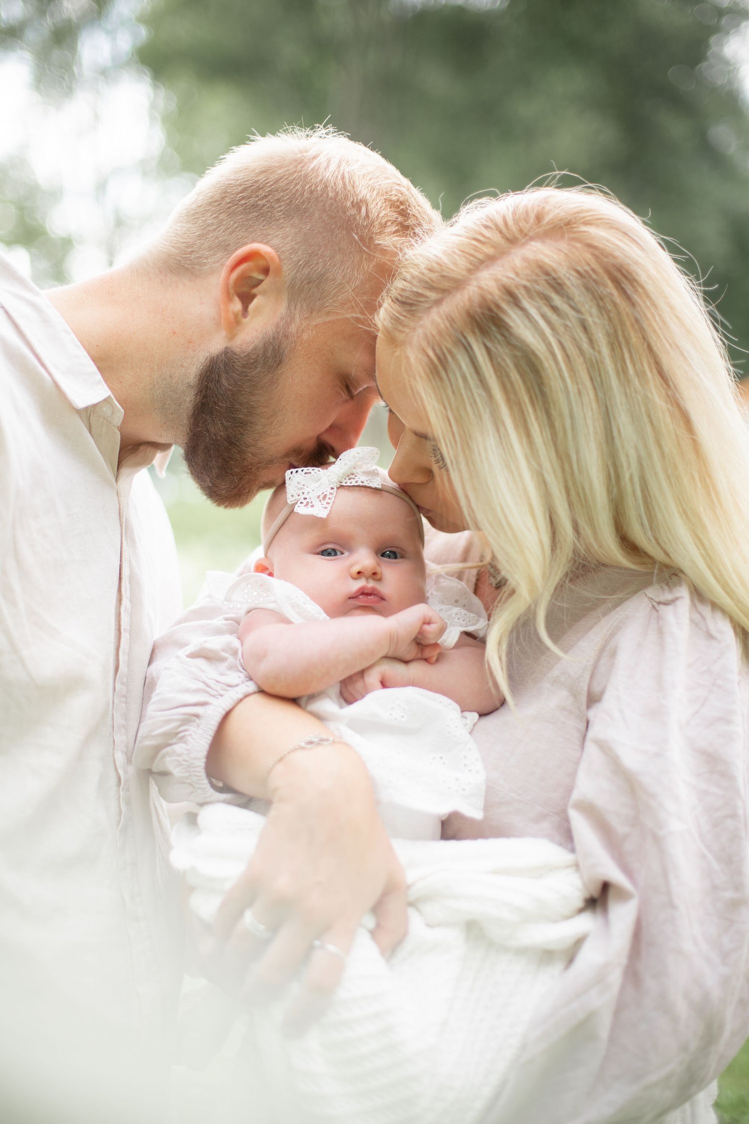 Mamma och pappa pussar nyfödd bebis på pannan. Nyföddfotografering utomhus i vacker sommargrön miljö. Familjefotograf i Katrineholm.