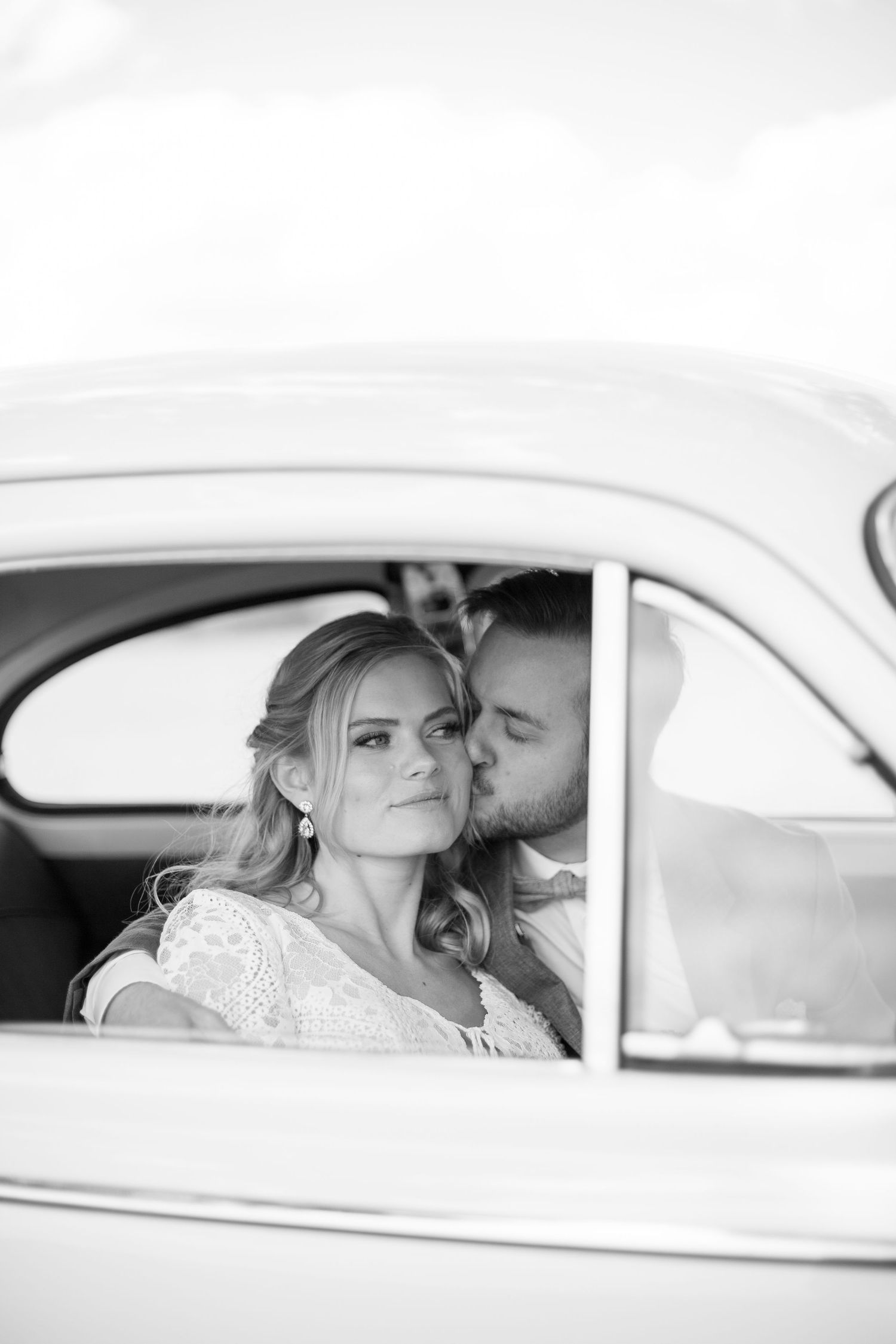 Vacker porträtt av brudpar under deras bröllopsfotografering. Sittandes i en bil, brudgummen kysser bruden på kinden. Bilden är svart/vit.