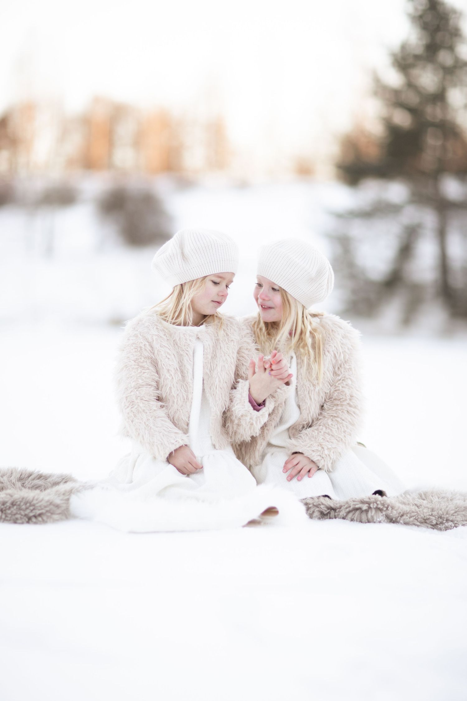 Barnfotografering på två systrar i matchande outfits utomhus i vackert snölandskap. Barnen håller i hand och lutar sig lite mot varandra.