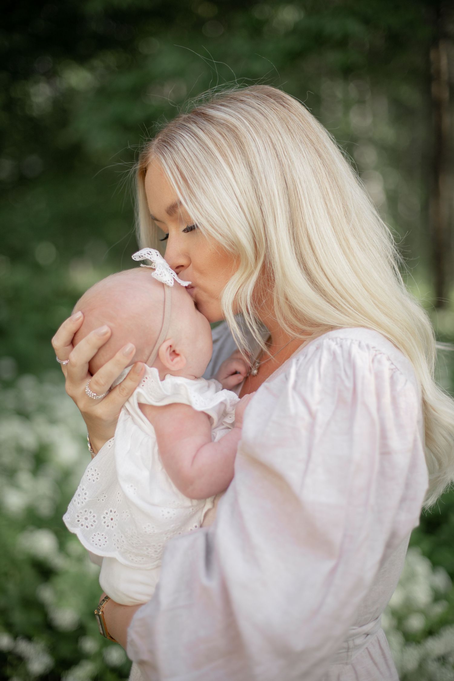 Mamma pussar sin nyfödda dotter på pannan. Nyföddfotografering utomhus i vacker sommargrön miljö. Familjefotograf i Katrineholm.
