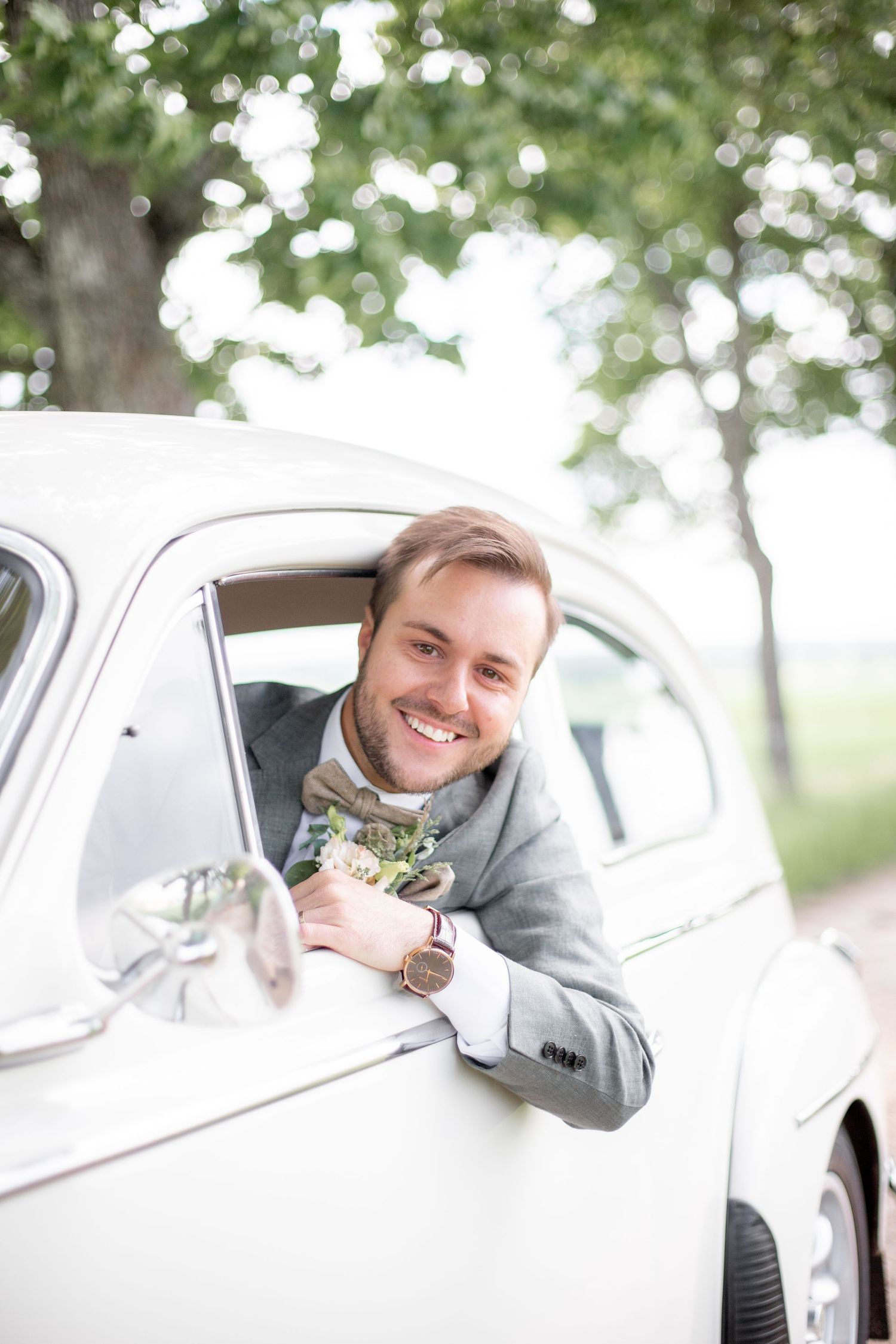 Brudgum hänger ut ur fönstret på en bil, lycklig under en bröllopsfotografering i somriga juni.