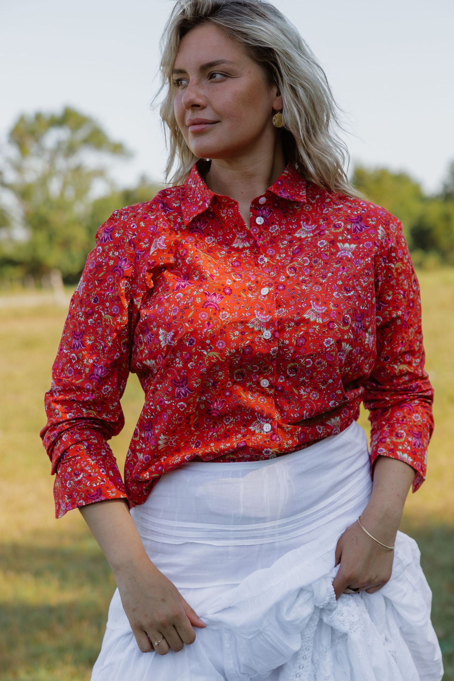 Woman wearing a red floral print button-up shirt with white skirt, standing outdoors in a green field.