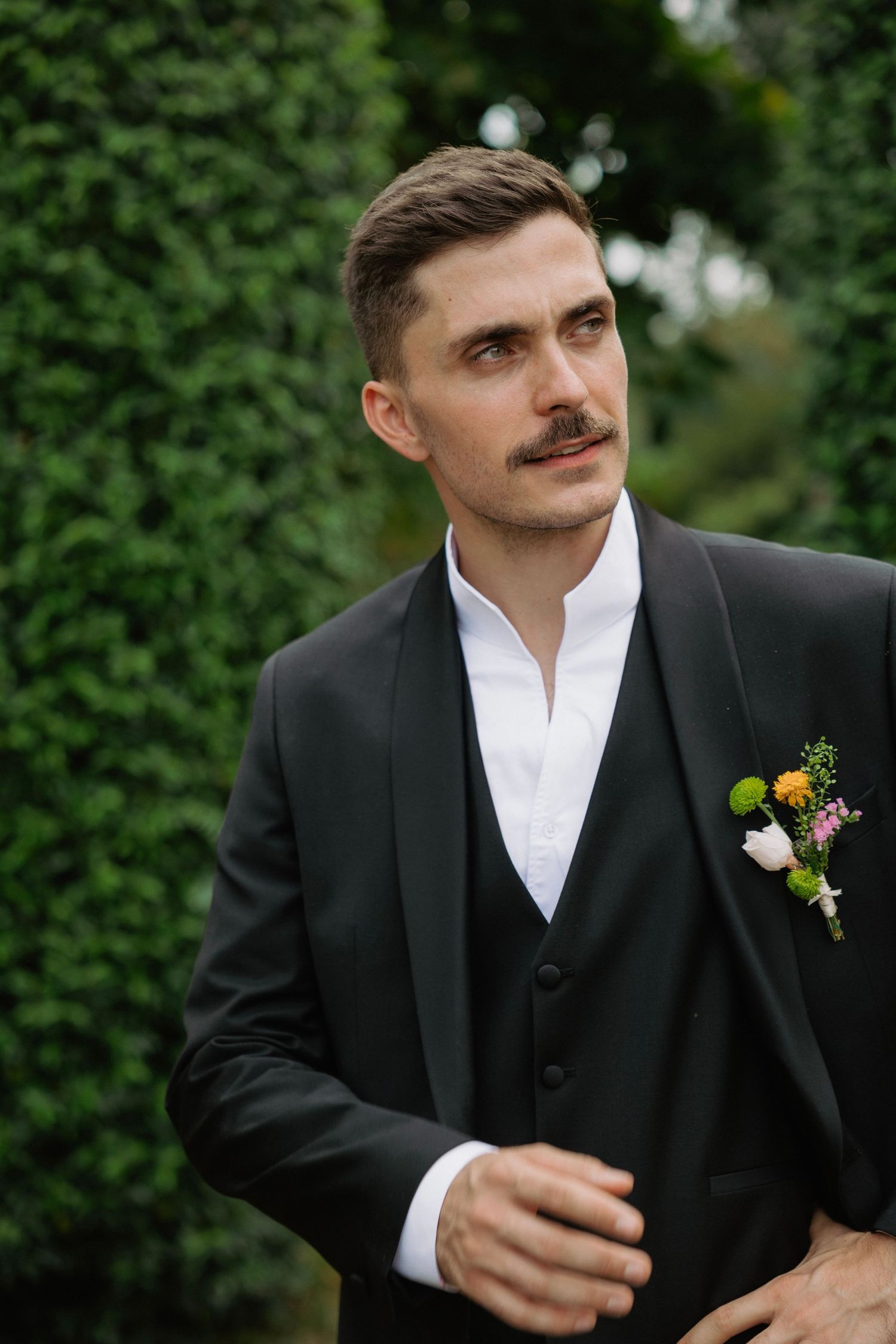 Elegant groom in black suit with floral boutonniere standing outdoors against lush green hedges.