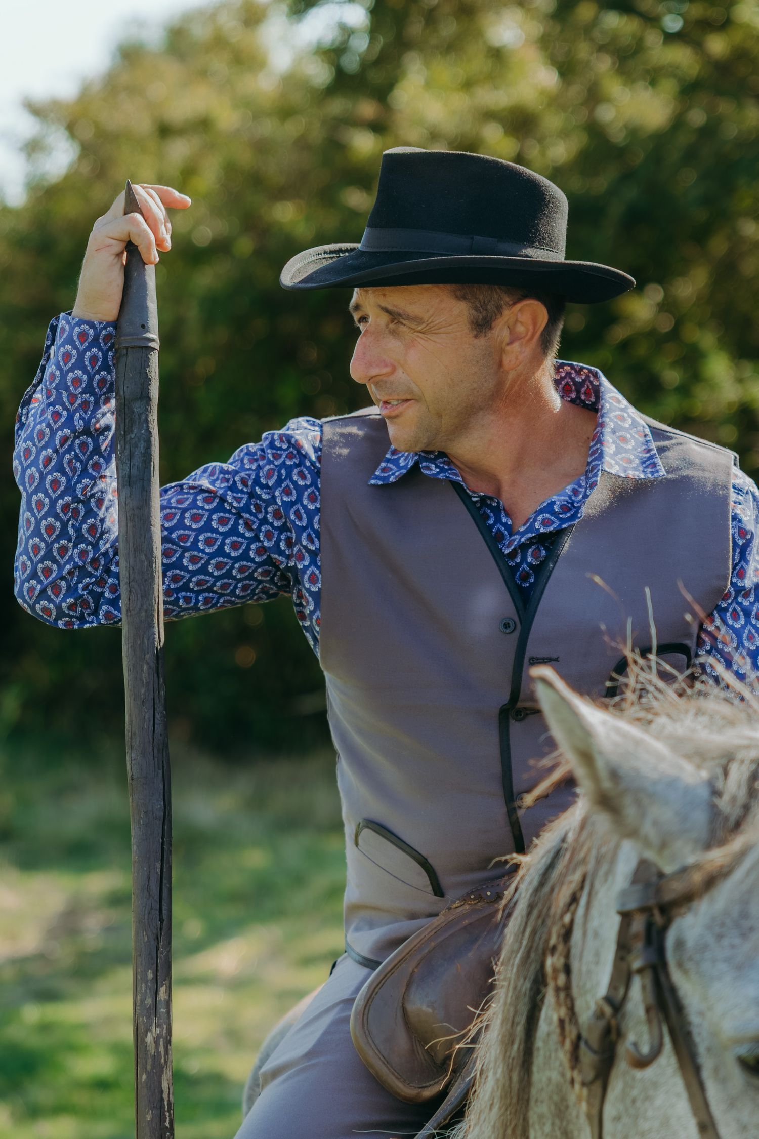A rider in western attire leans against a wooden fence post in a rural setting.