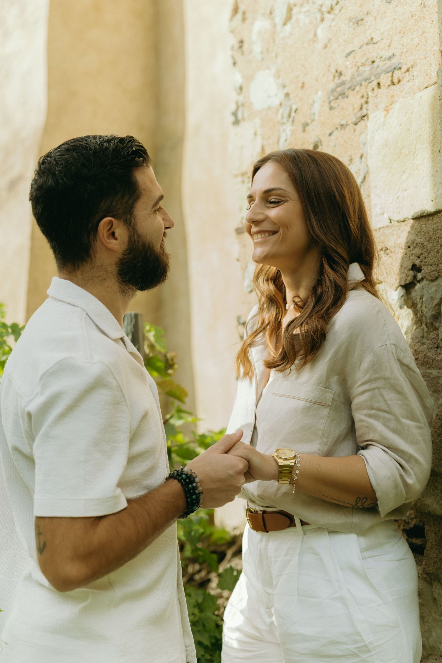 A couple sharing a joyful moment while wearing coordinated white outfits against a stone wall.