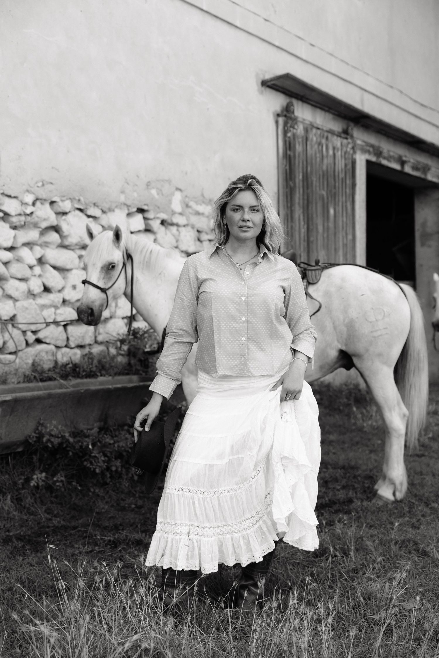 Black and white photo of a blonde woman in a white skirt and blouse standing beside a white horse near a stone barn.