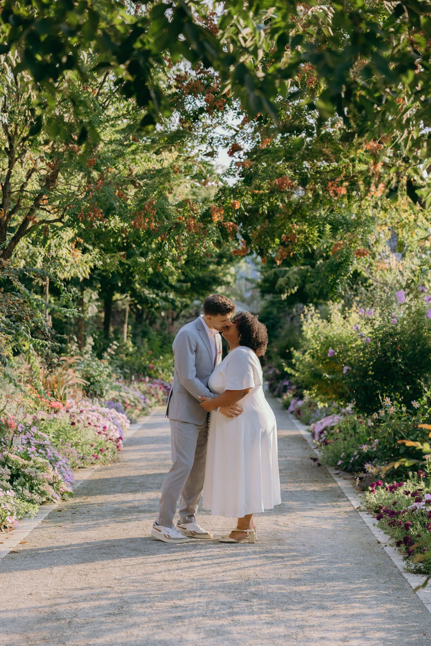 A couple embraces on a garden path surrounded by blooming flowers and greenery.