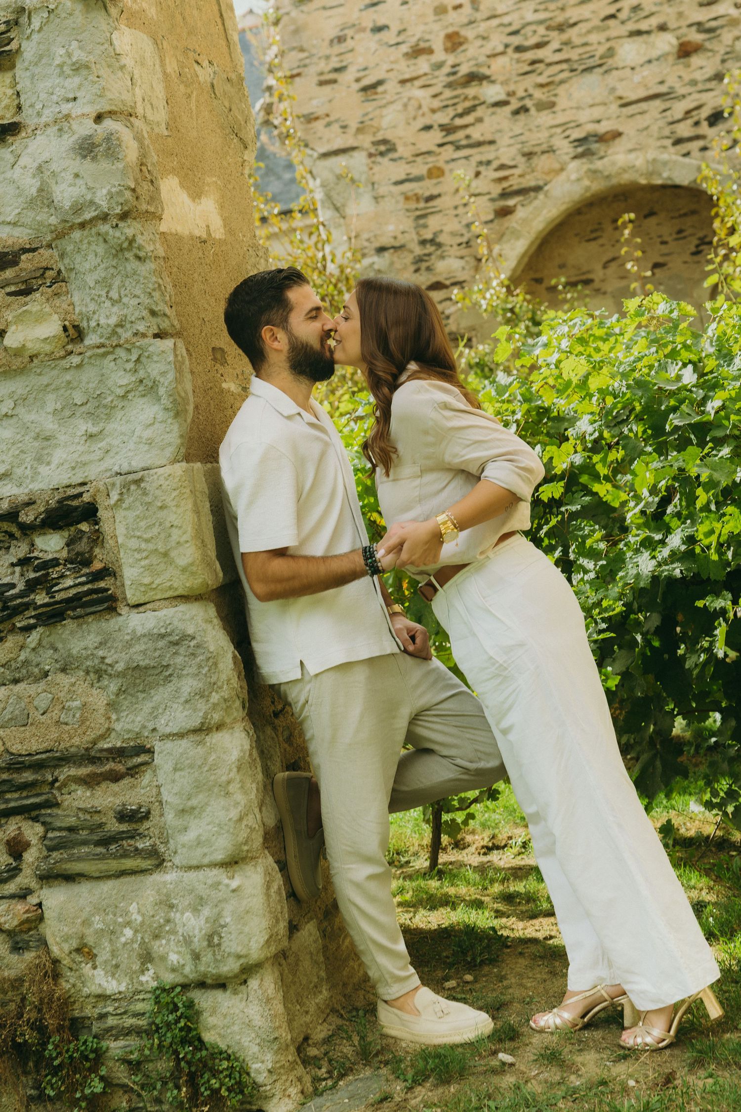 A couple shares a romantic moment against a rustic stone wall surrounded by green foliage on a sunny day.