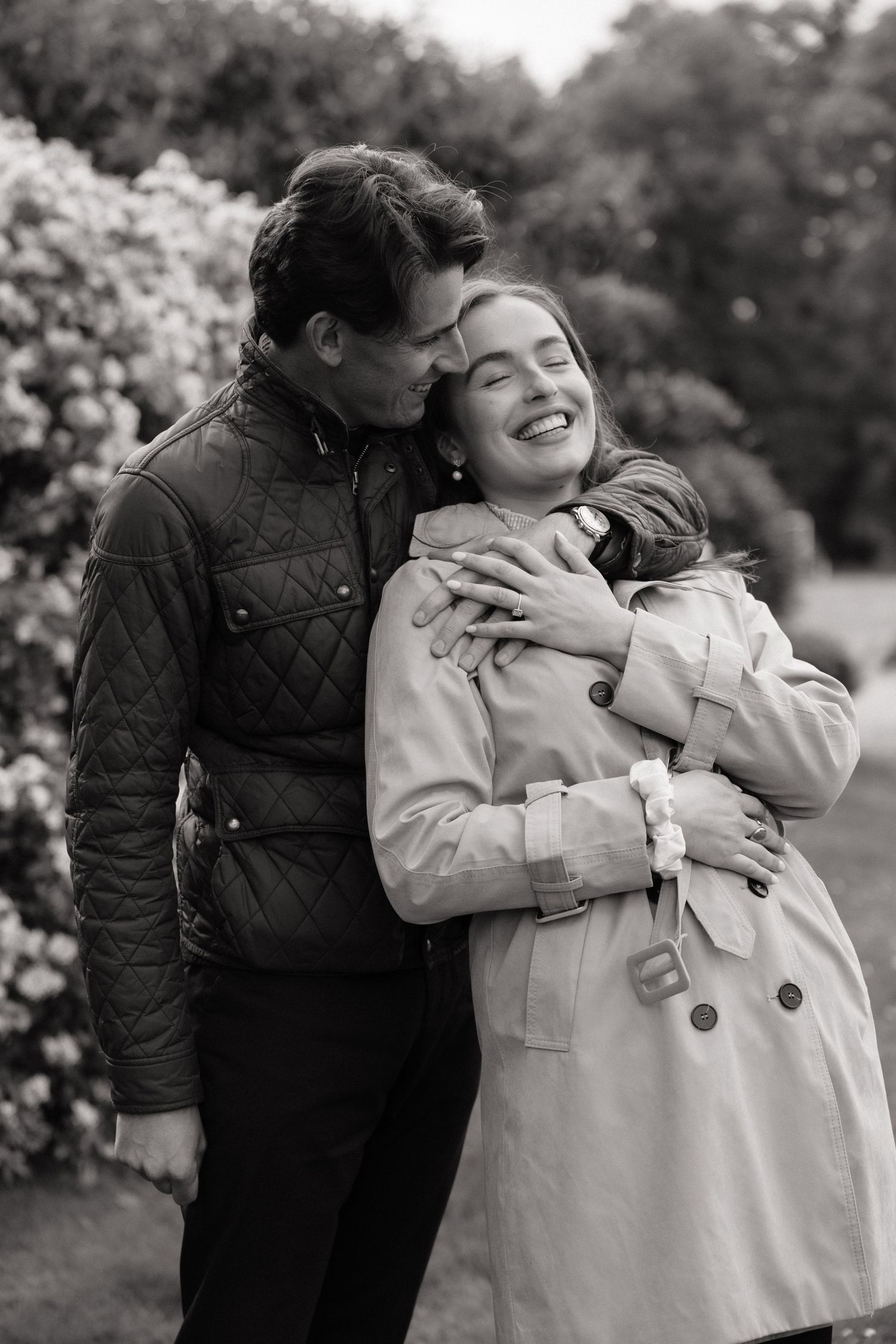 Black and white photo of a couple embracing in a garden, man kissing womans head as she smiles joyfully.