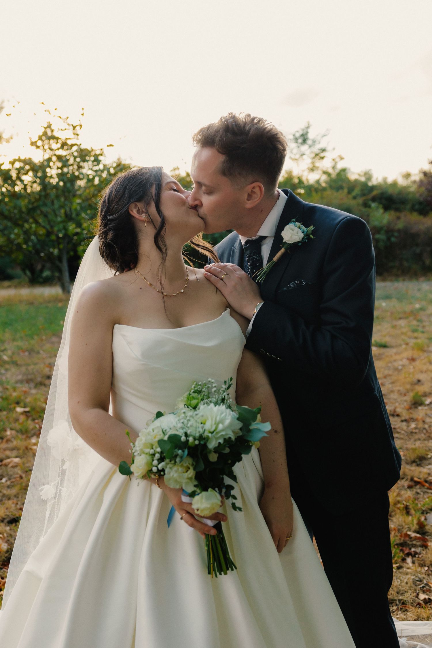 Bride and groom sharing a kiss outdoors, bride holding white floral bouquet in elegant strapless gown with veil.