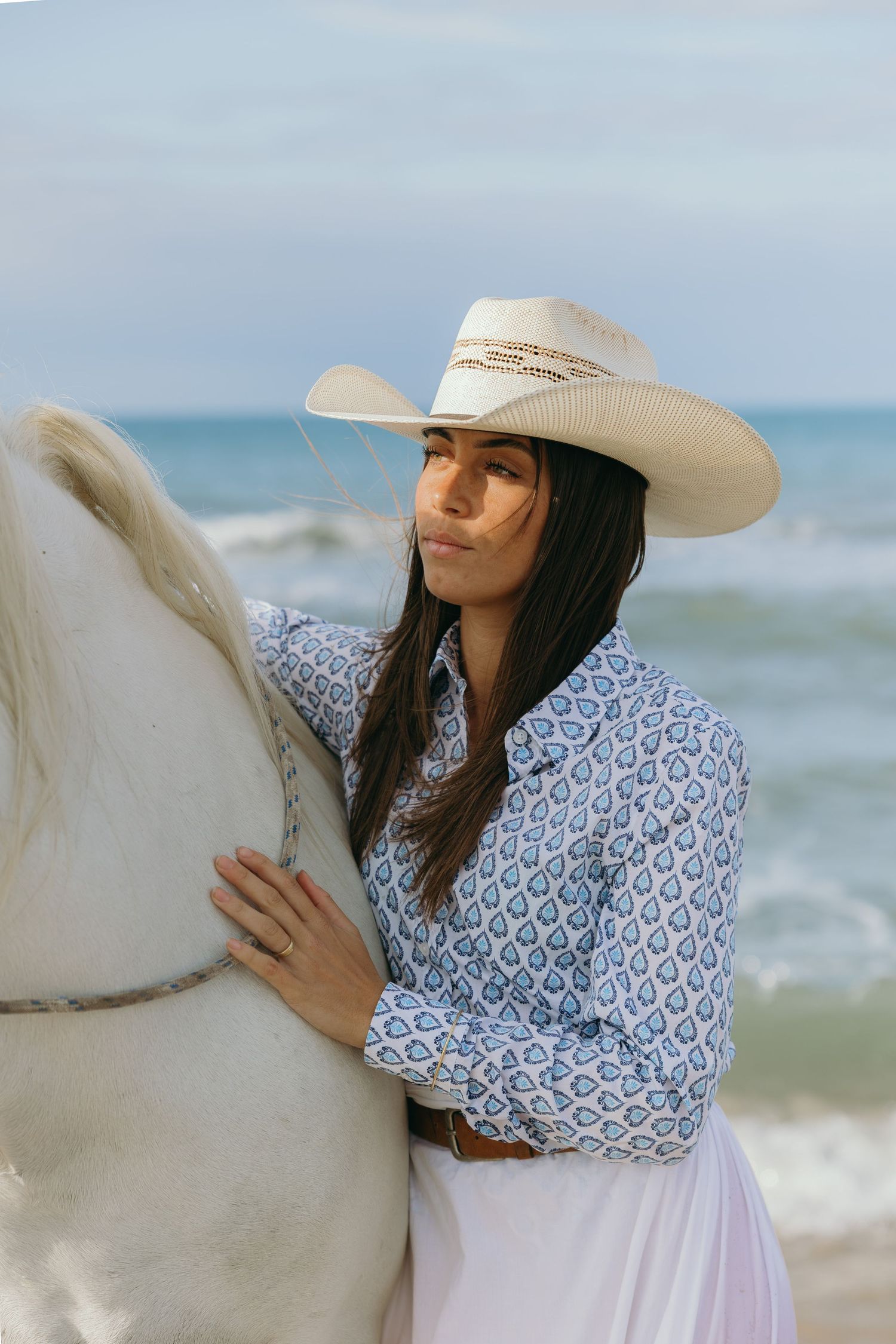 A person wearing a white cowboy hat and patterned dress embraces a white horse by the ocean.