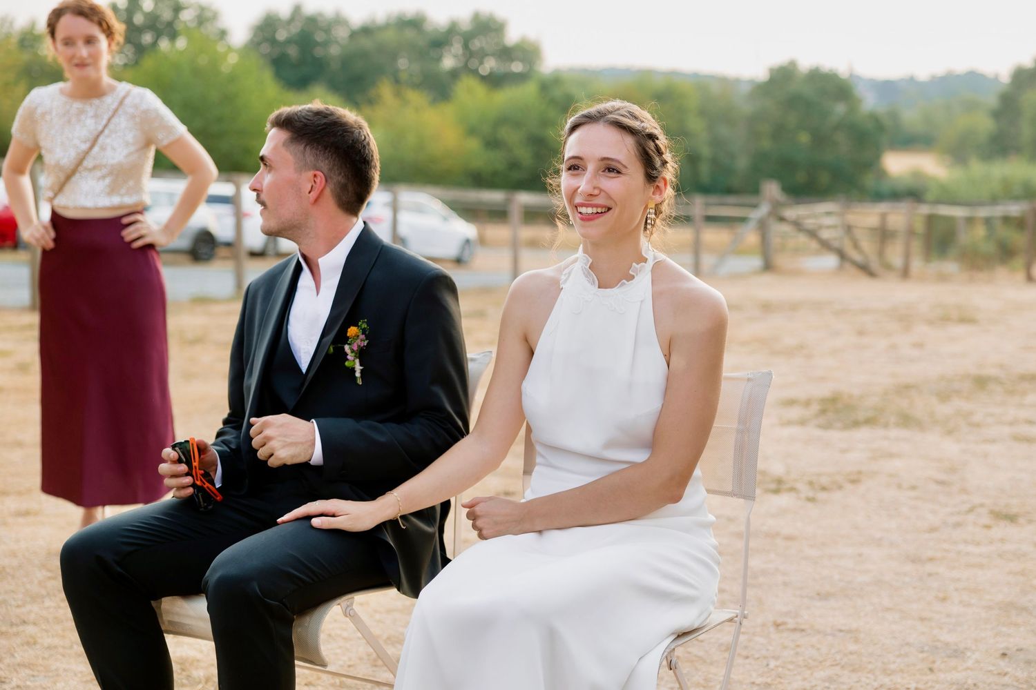 Bride in white halter dress and groom in black suit sit together outdoors at a rustic wedding venue.