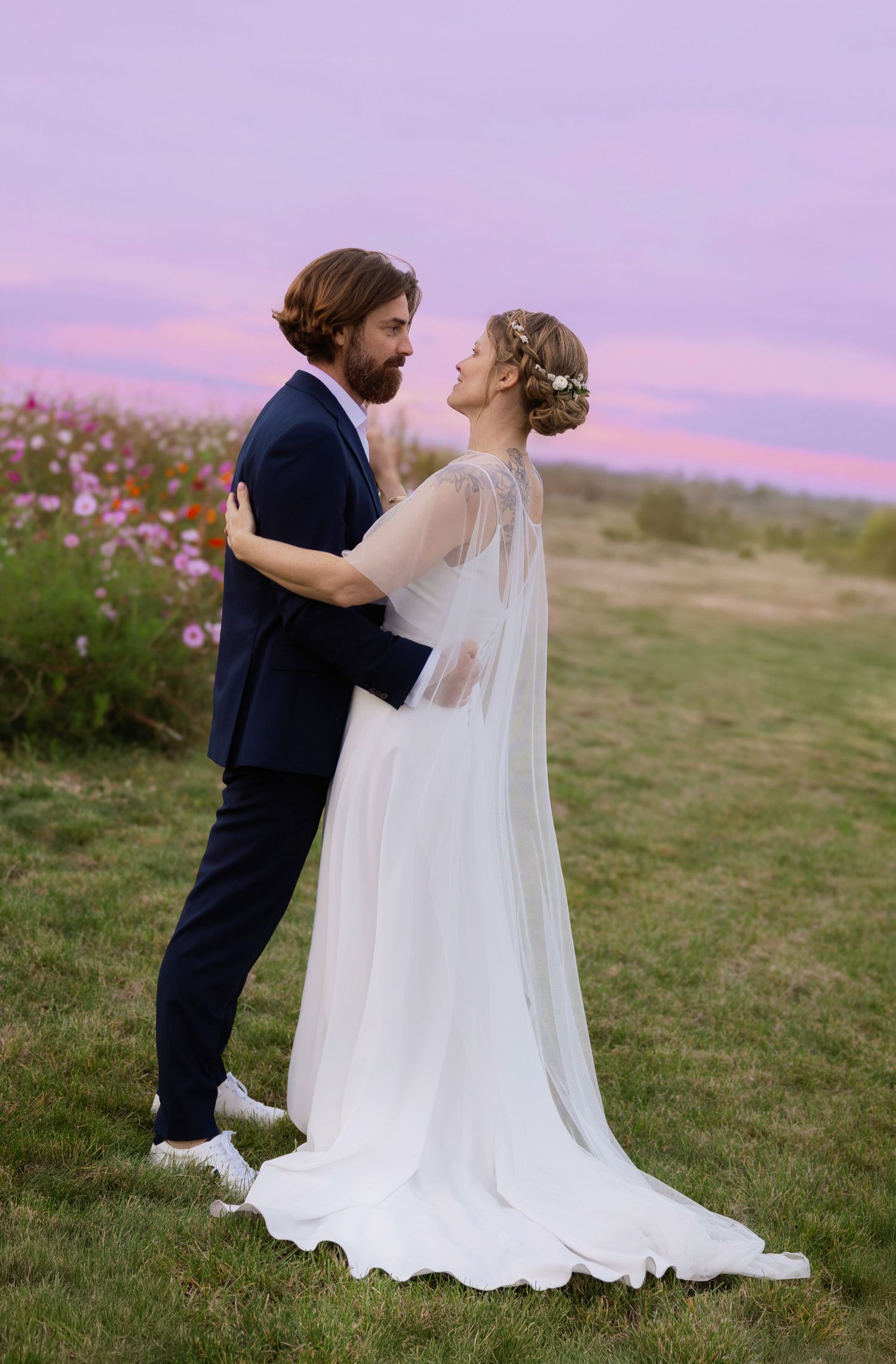 A couple in wedding attire share a romantic moment in a field at sunset with a flowing white veil and navy suit.