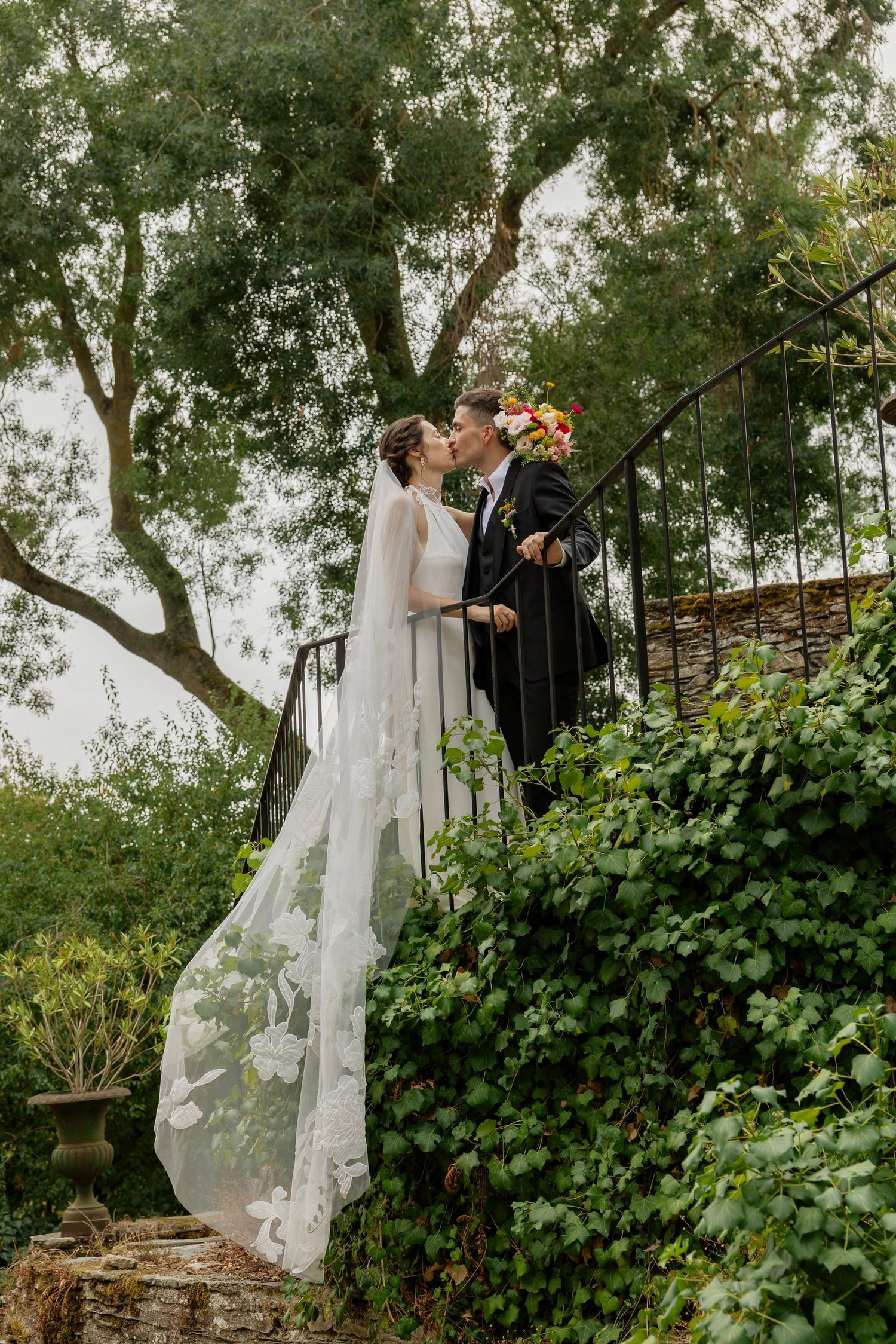 Bride and groom share a kiss on garden stairs, surrounded by lush greenery, with a flowing lace wedding veil.
