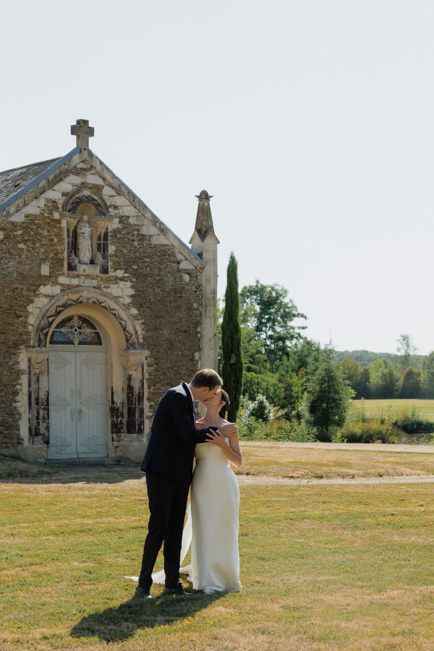 Bride and groom share a romantic kiss in front of a historic stone chapel surrounded by lush greenery on their wedding day.
