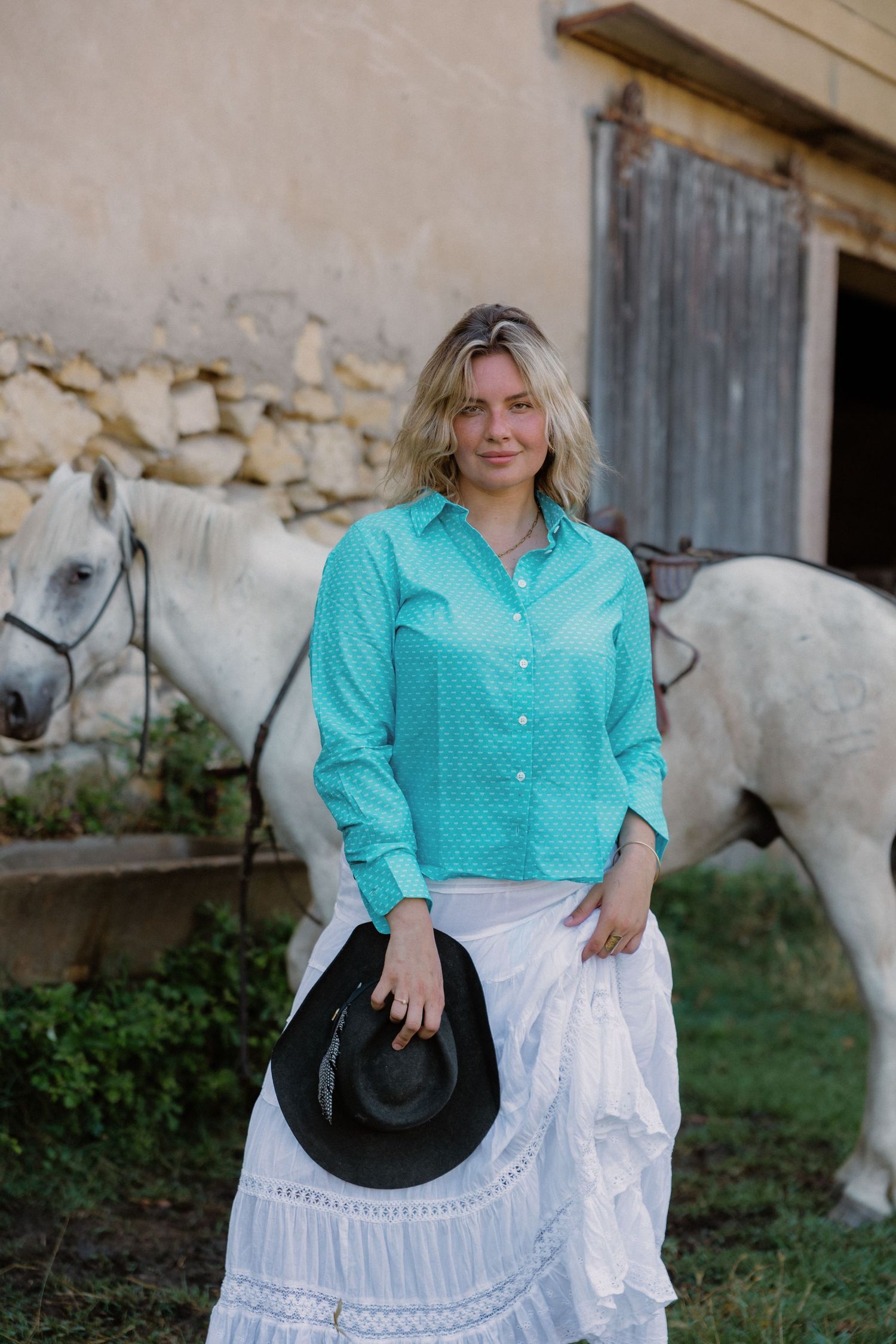 Woman in turquoise shirt and white skirt holding black cowboy hat, standing near a white horse outside a stable.