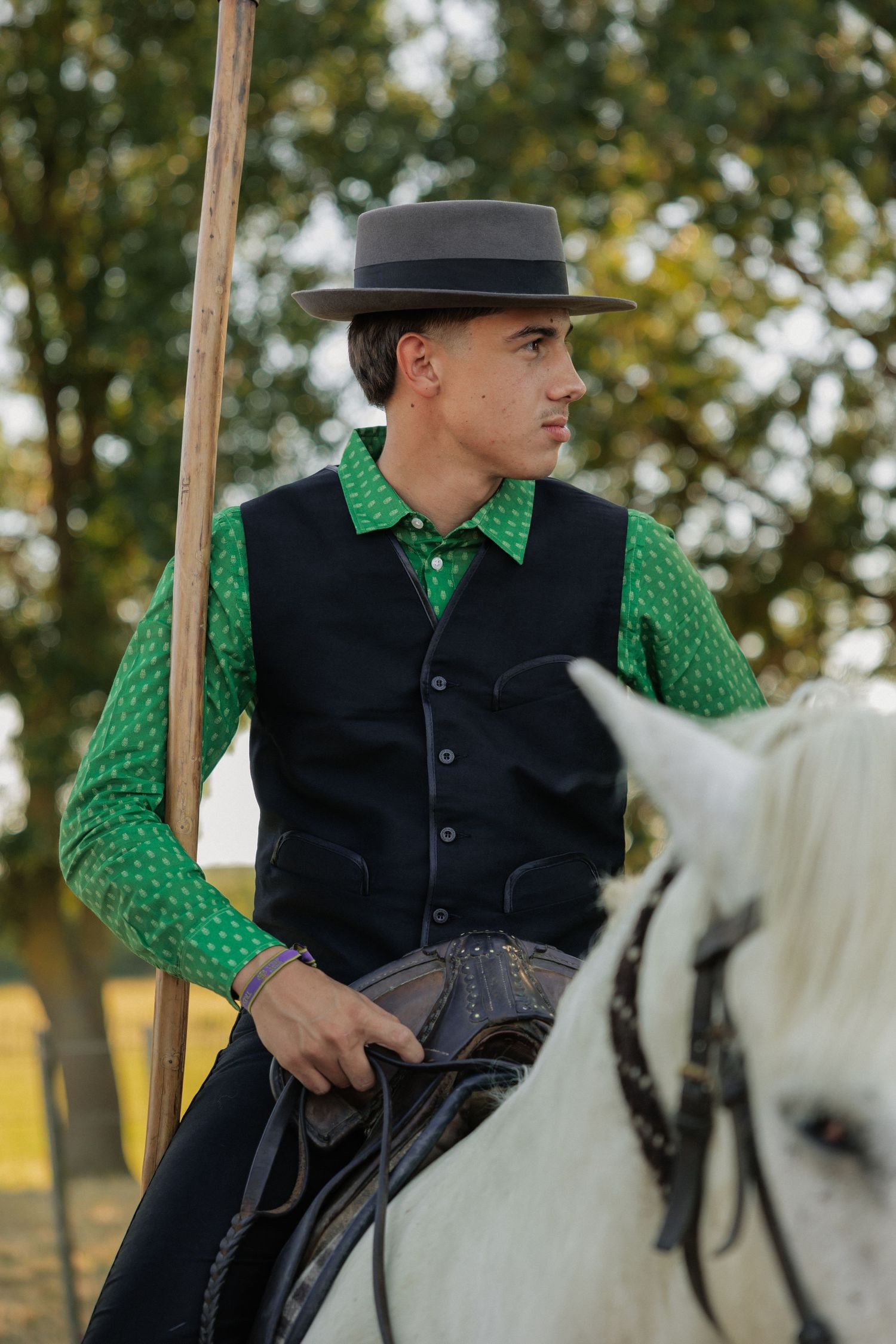 Young man in green shirt, black vest, and wide-brim hat riding a white horse outdoors.