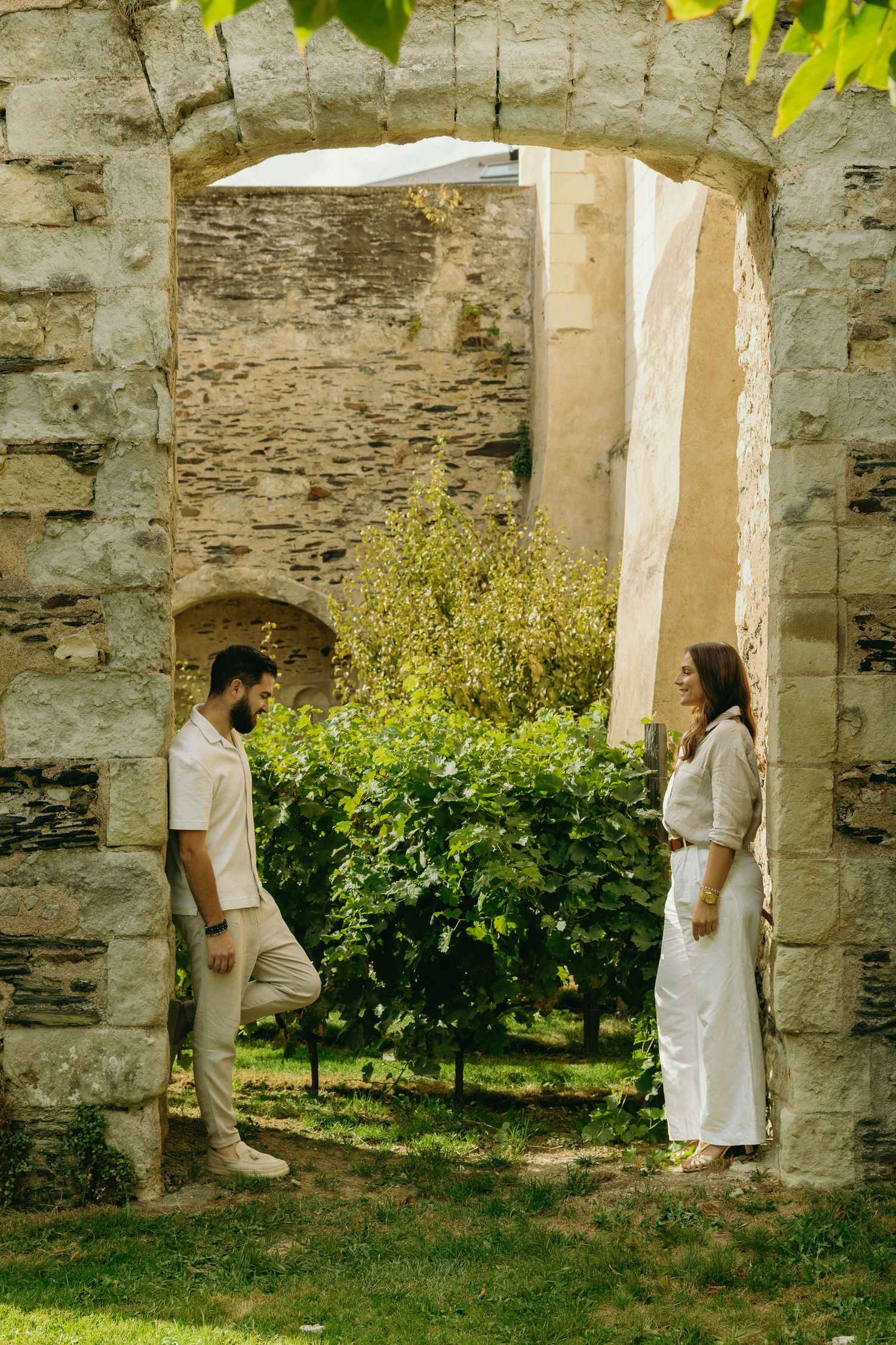 A couple in all-white outfits standing in a stone archway surrounded by greenery.