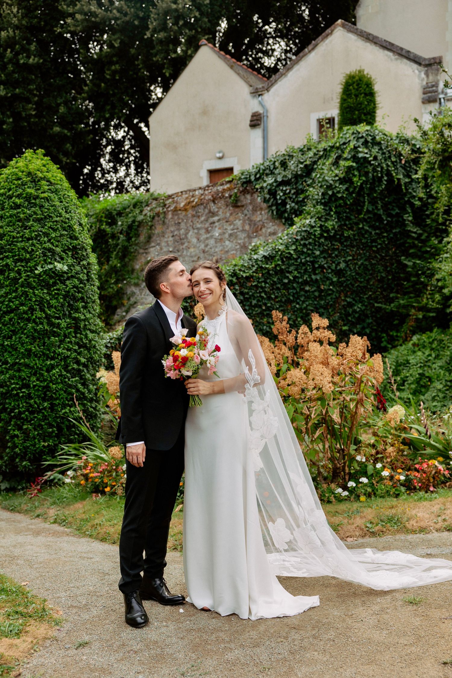 Groom kissing bride on cheek in lush garden, bride holding colorful bouquet with long veil.