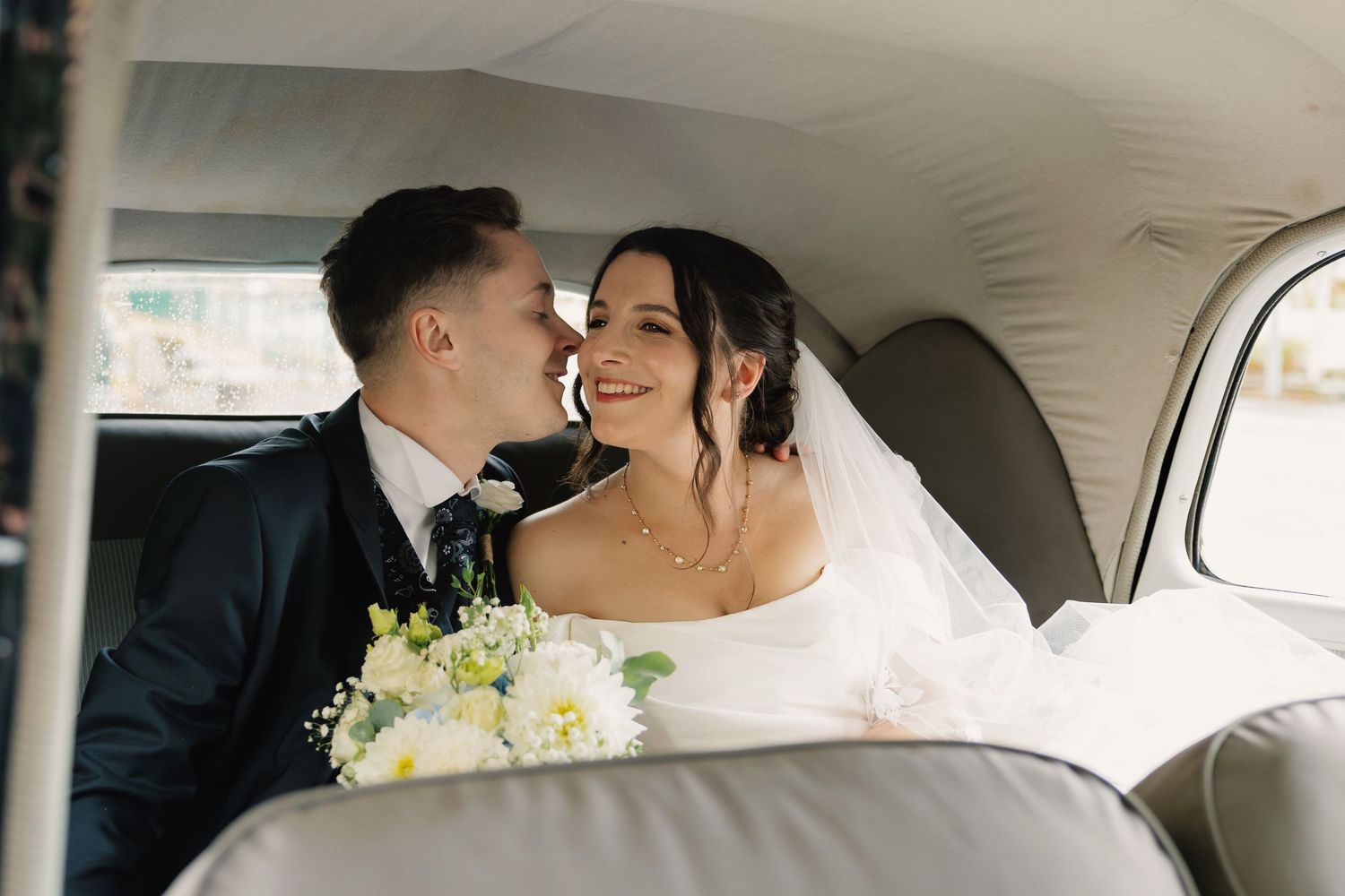 Bride and groom sharing a romantic moment inside a vintage wedding car, bride holding a white bouquet.