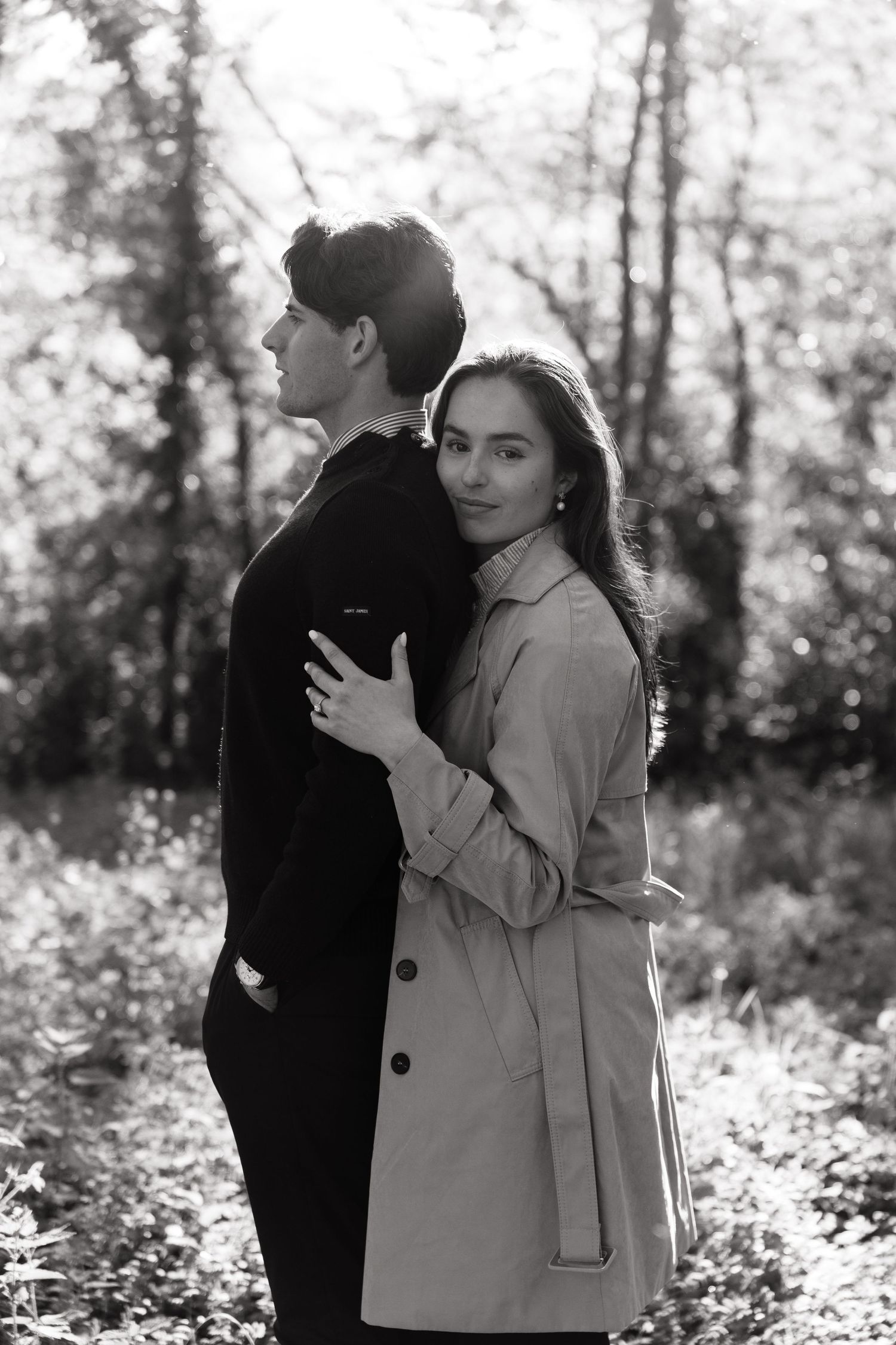 Black and white photo of a young couple embracing in a forest, woman in trench coat smiling at camera.