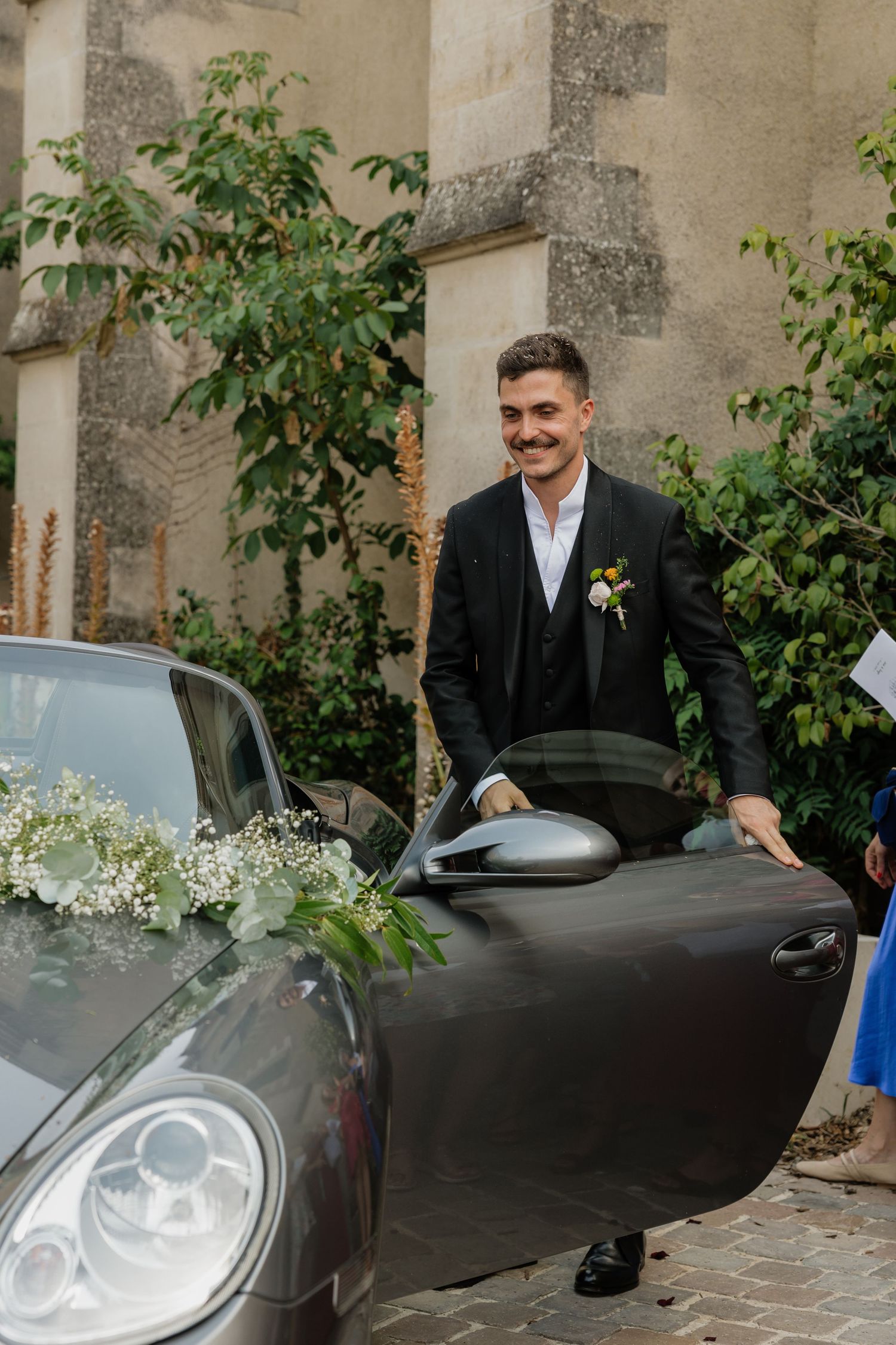 Smiling groom in black suit with boutonniere opening door of flower-decorated car at wedding venue.