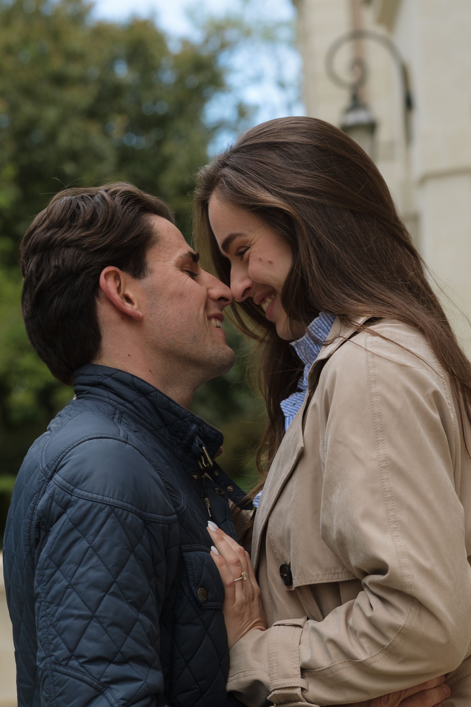 A smiling couple sharing a romantic moment outdoors, nearly kissing, wearing casual jackets with greenery in the background.