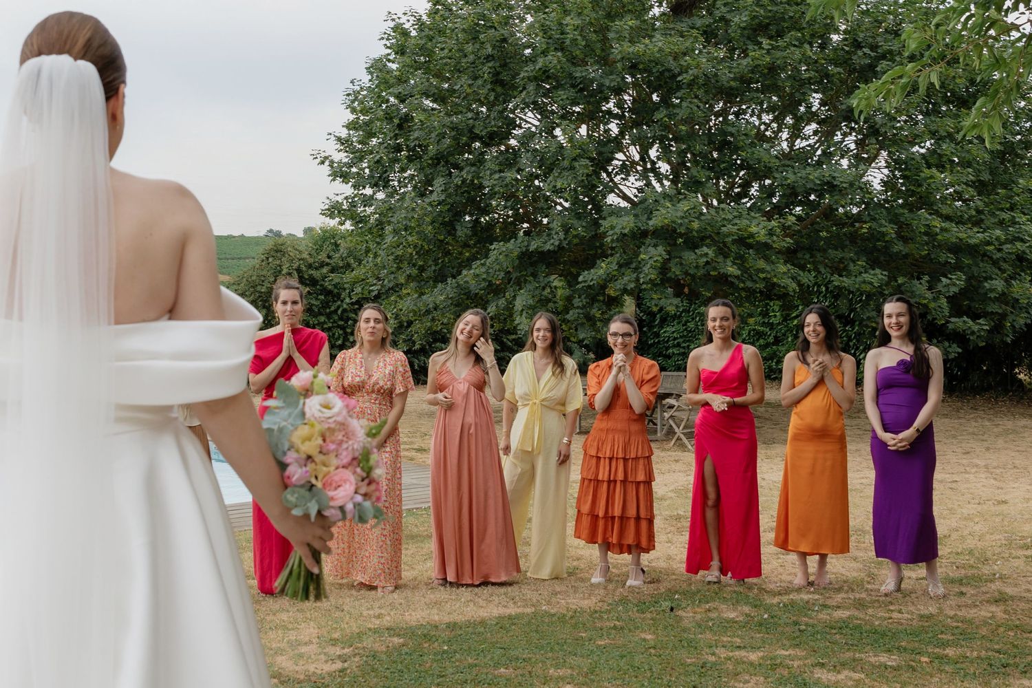 Bride faces colorful bridesmaids in mismatched rainbow dresses standing in a line outdoors.