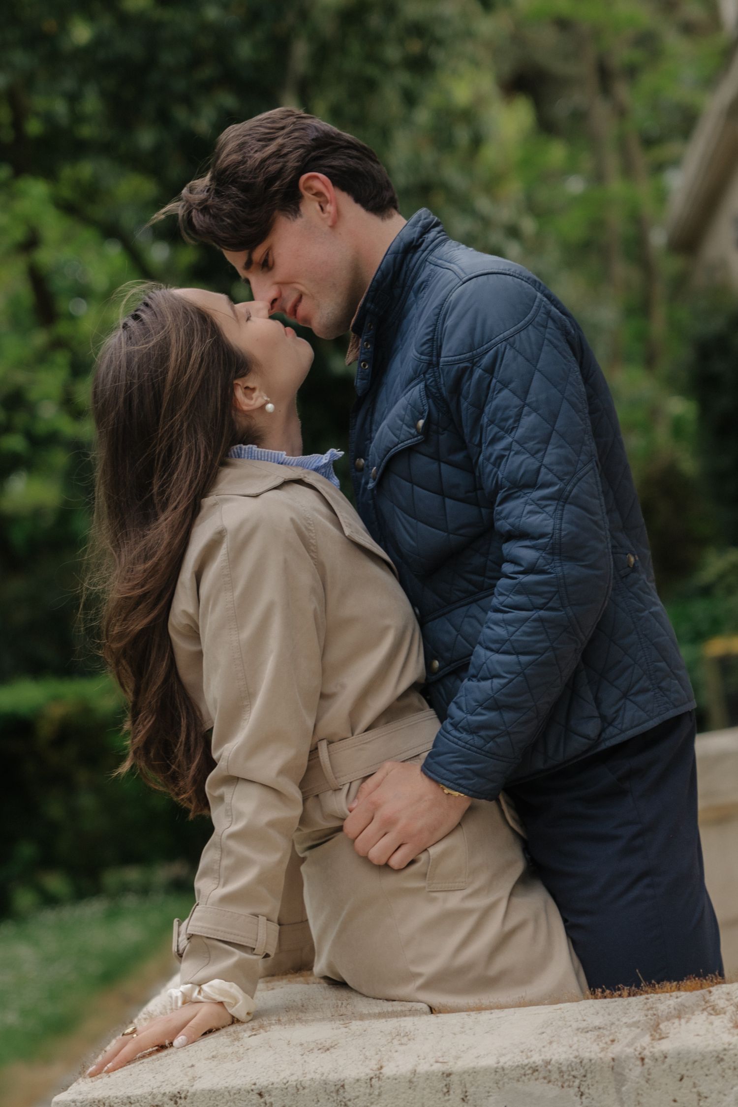 Romantic couple sharing a kiss outdoors, woman in beige trench coat and man in navy quilted jacket, green trees background.