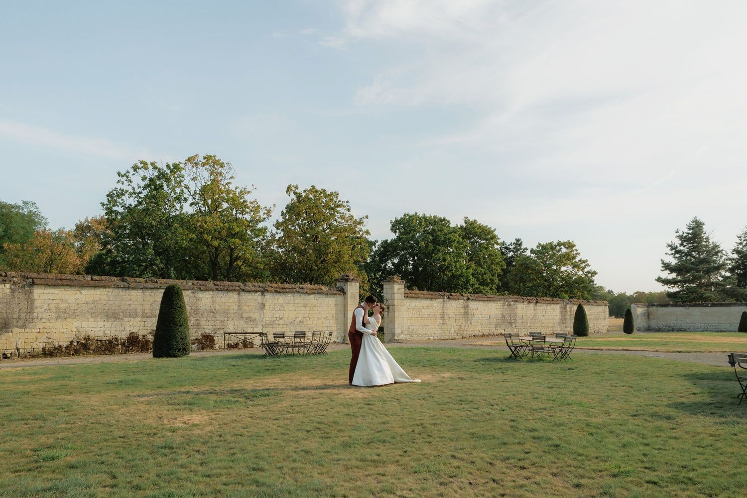 Bride and groom embrace in a sunlit garden courtyard with stone walls and manicured grounds at a French chateau wedding.